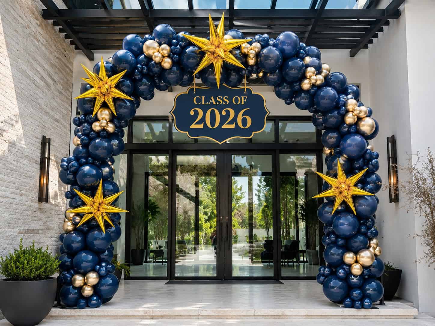 A balloon arch with blue and gold balloons and star decorations frames an entrance with a “Class of 2026” sign above glass doors.