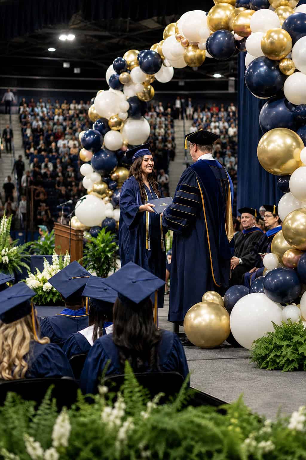 A graduate in a blue cap and gown receives her diploma on stage during a commencement ceremony, with an audience and balloon arch in the background.