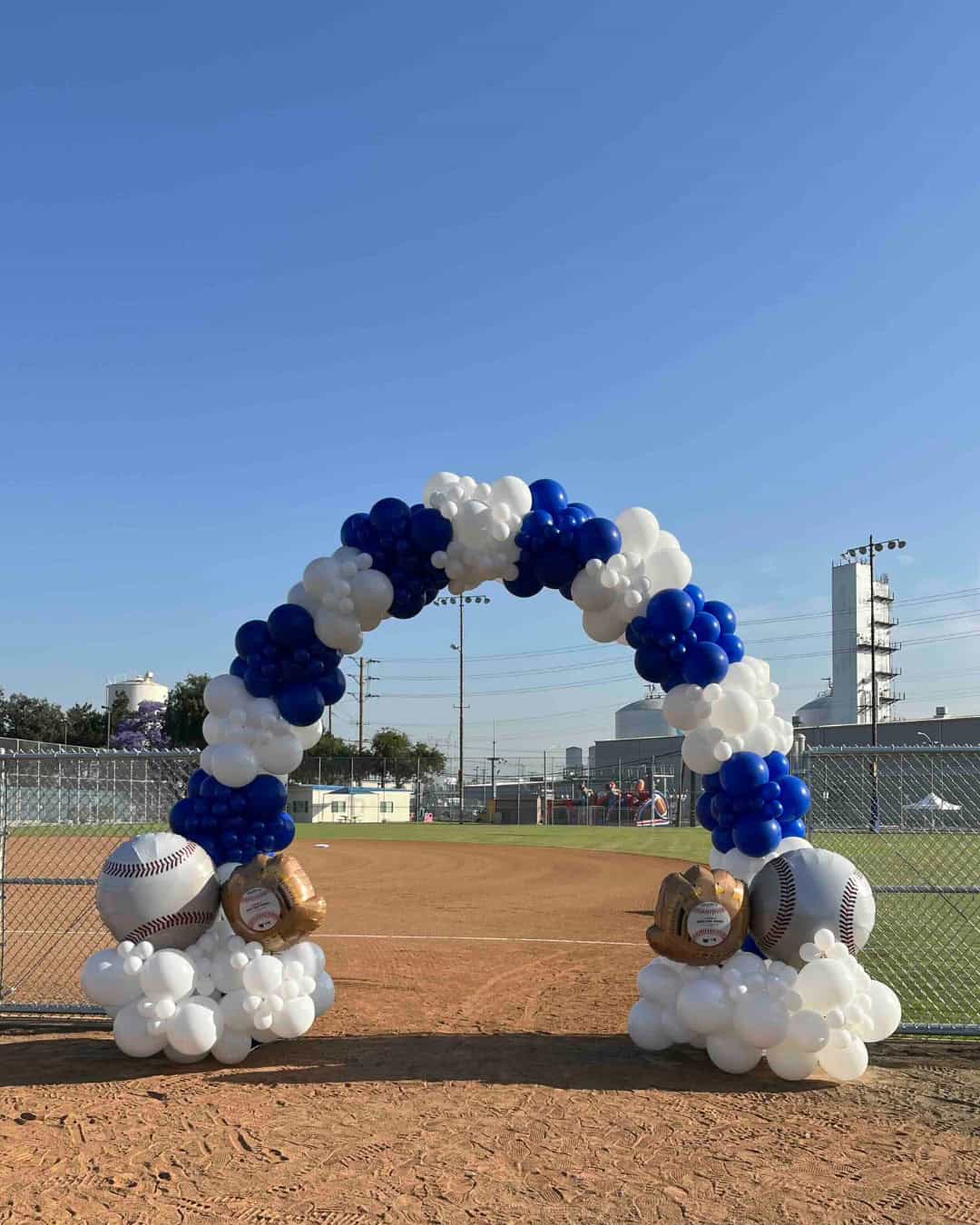 A balloon arch decorated with blue, white, and brown balloons, shaped like baseballs and gloves, stands on a baseball field near a chain-link fence—perfect for sports events or celebrations.