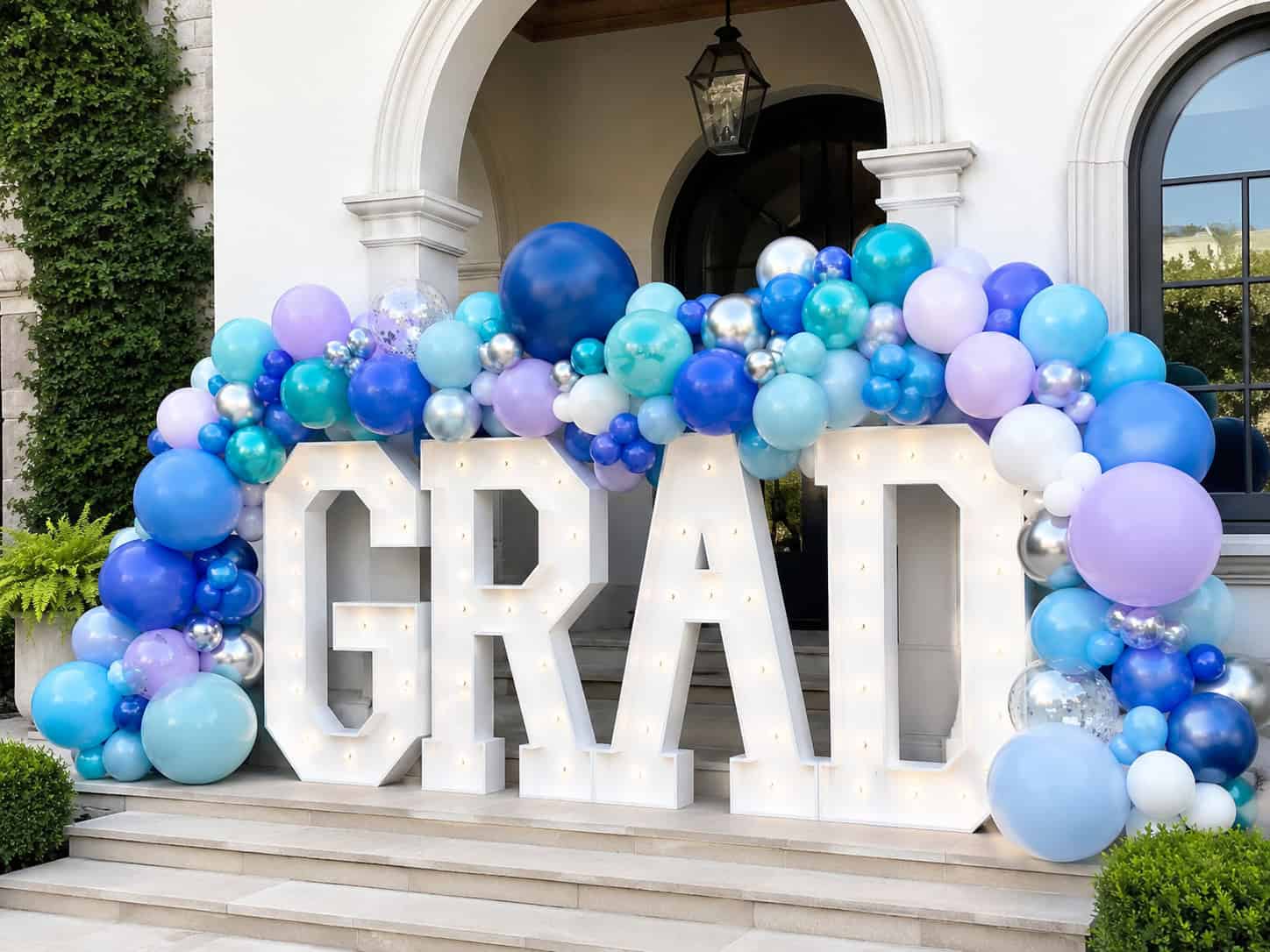 Large light-up letters spelling "GRAD" are decorated with a blue, purple, and white balloon arch, positioned in front of building steps and an arched doorway.