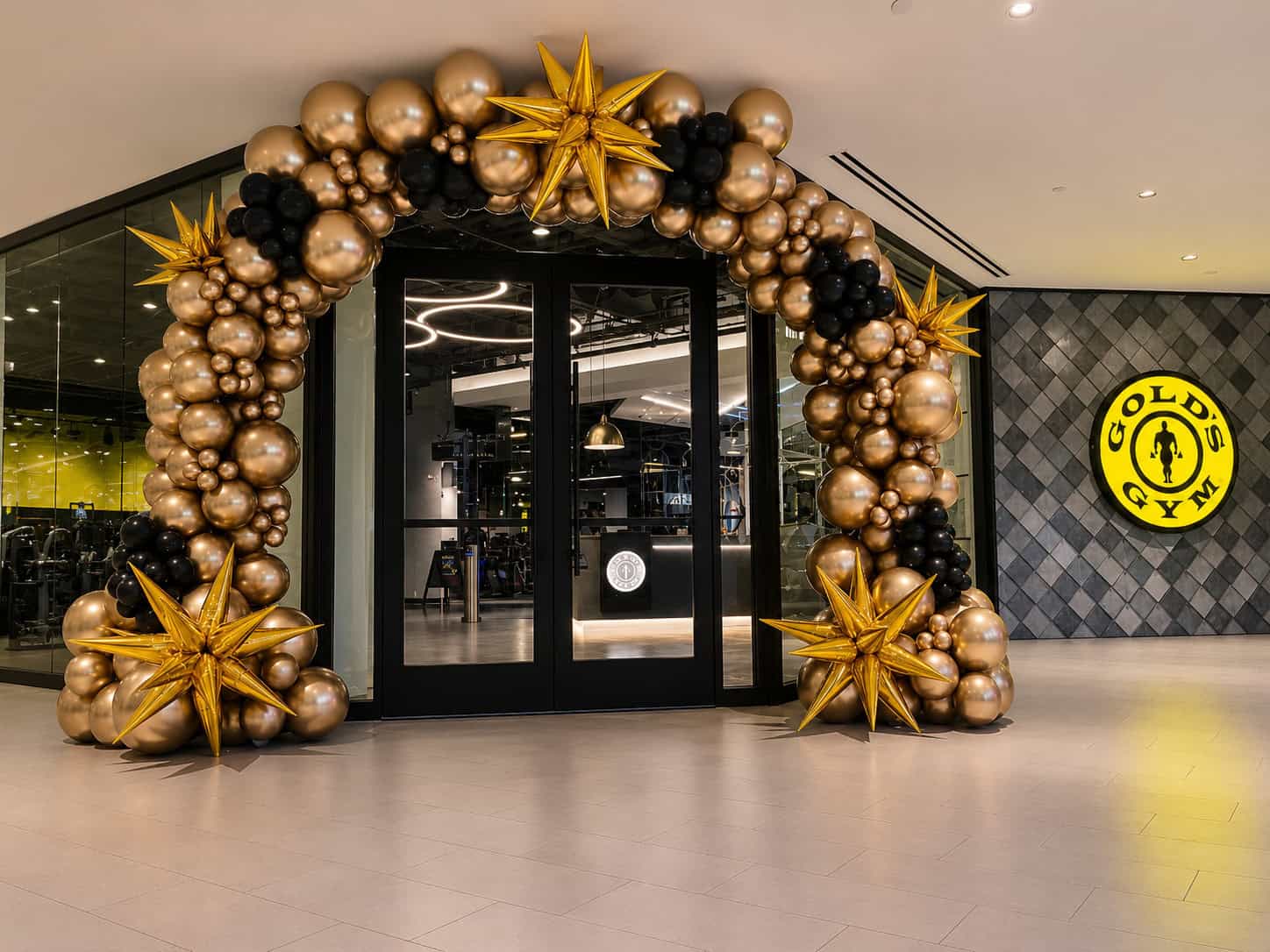 Gold’s Gym entrance decorated with gold and black balloons and gold star-shaped ornaments, celebrating Grand Openings with the gym’s logo visible on the wall.