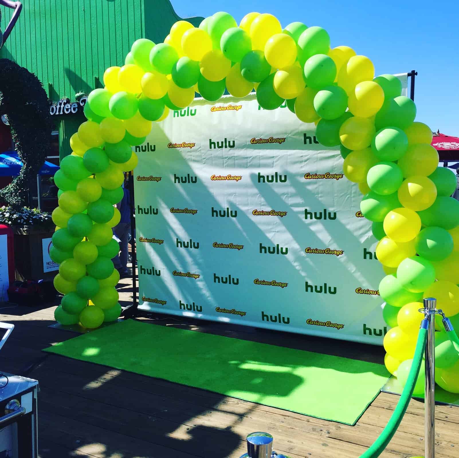 A green and yellow balloon arch, part of a vibrant display of balloon arches, stands in front of a step-and-repeat banner featuring the Hulu and "Casual Sweep" logos on a sunny day.