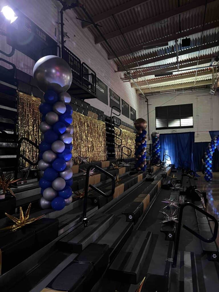 Decorated gym bleachers with blue and silver balloon columns, gold tinsel backdrop, metallic star decorations, and blue drapery in the background—perfect for a TV production or film production setting.