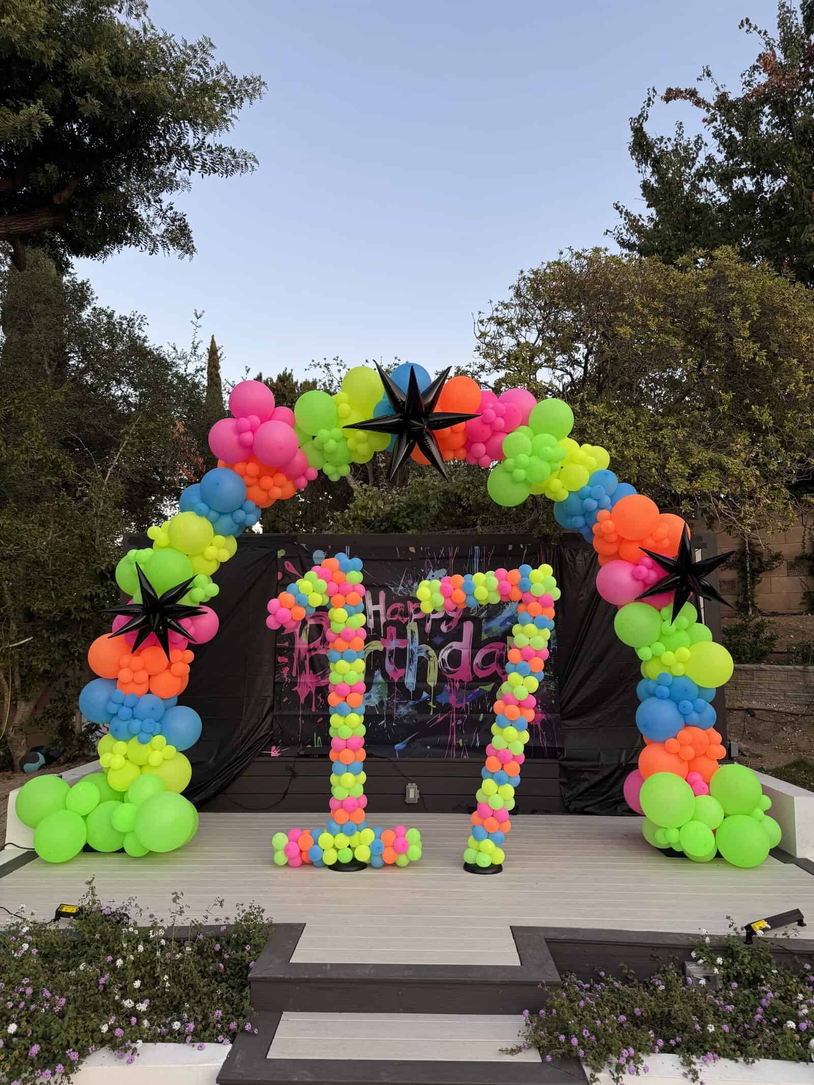 A colorful balloon arch and large "17" decoration stand on an outdoor stage, with a "Happy Birthday" sign in the background.