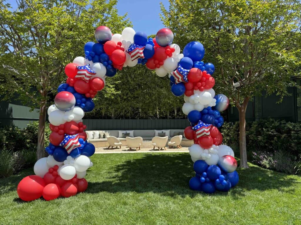 A festive balloon arch in red, white, and blue with small American flag designs stands on a lawn between two trees, with outdoor seating visible in the background—perfect for celebrating with patriotic balloon arches.