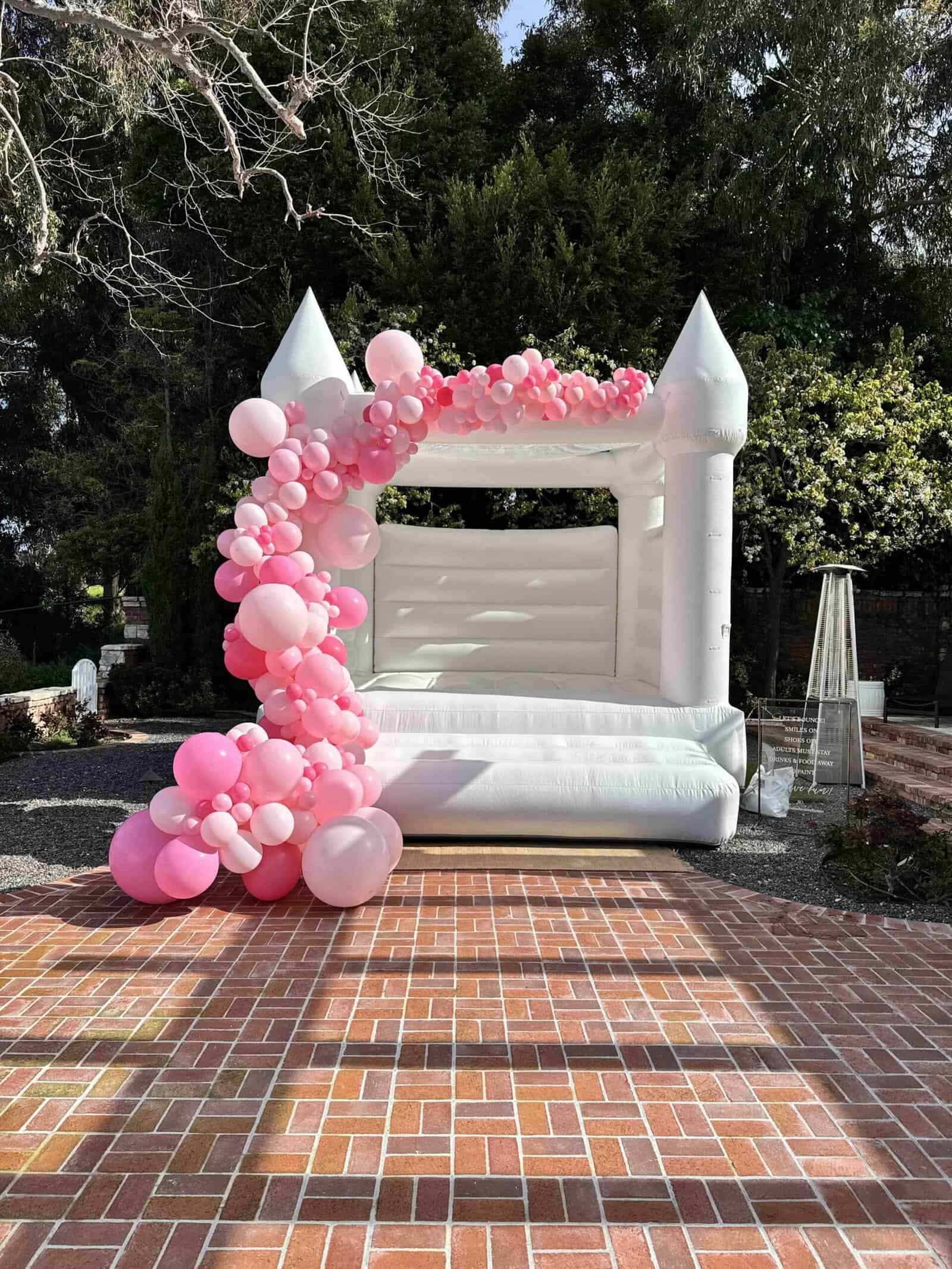 A white inflatable bounce house adorned with pink and white balloon garlands stands on a brick patio outdoors, offering charming party decor amid trees and greenery in the background.
