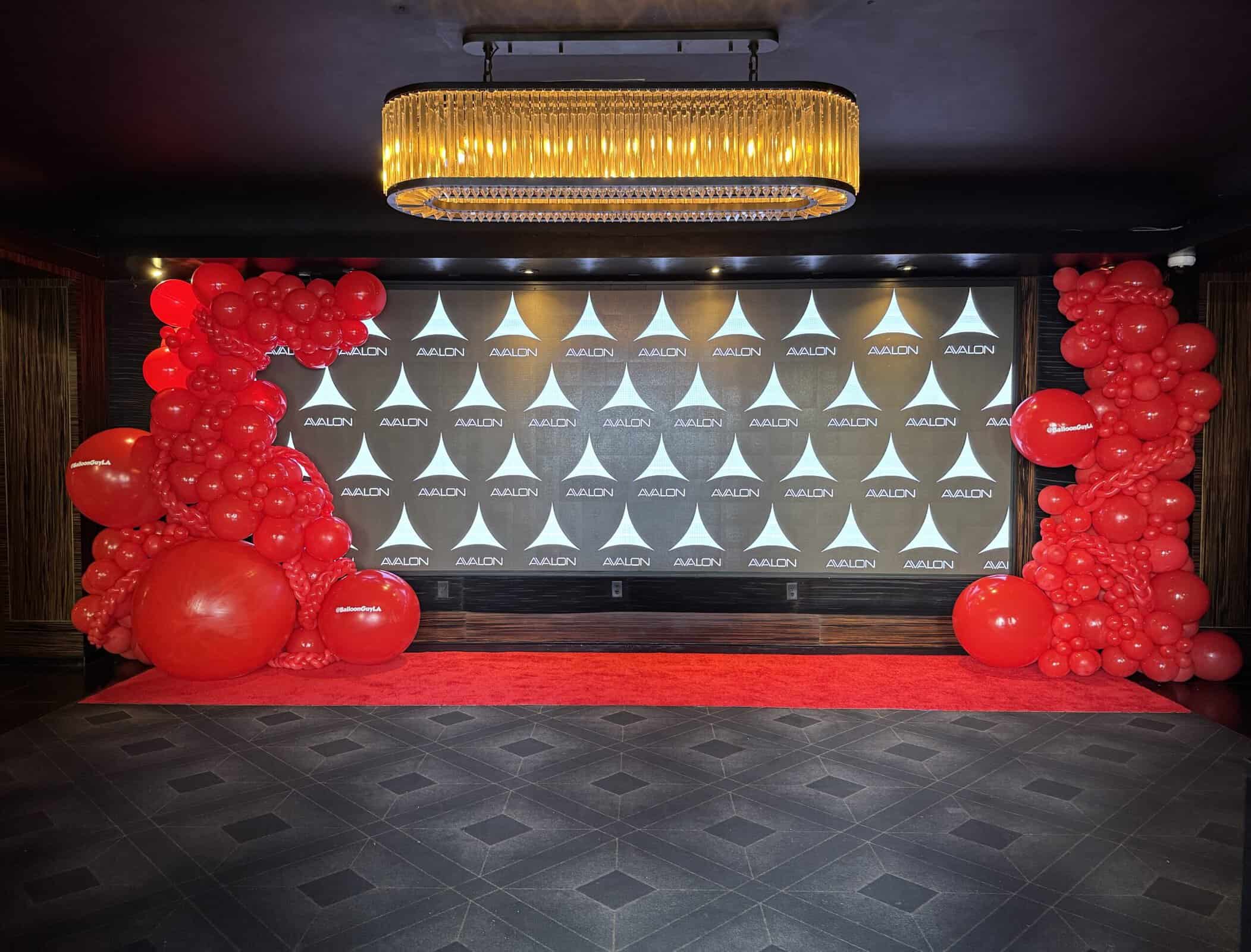 A decorated event backdrop with a red carpet, clusters of red balloons on both sides, and a repeating “ALOTN” logo pattern under a large rectangular chandelier—perfect for memorable Corporate Events.