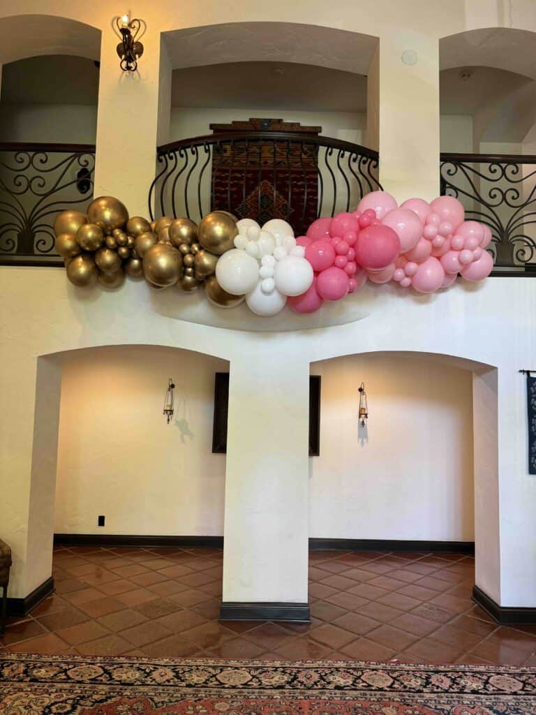 A garland of gold, white, and pink balloon decorations is displayed on the balcony railing above an arched hallway in an indoor space.