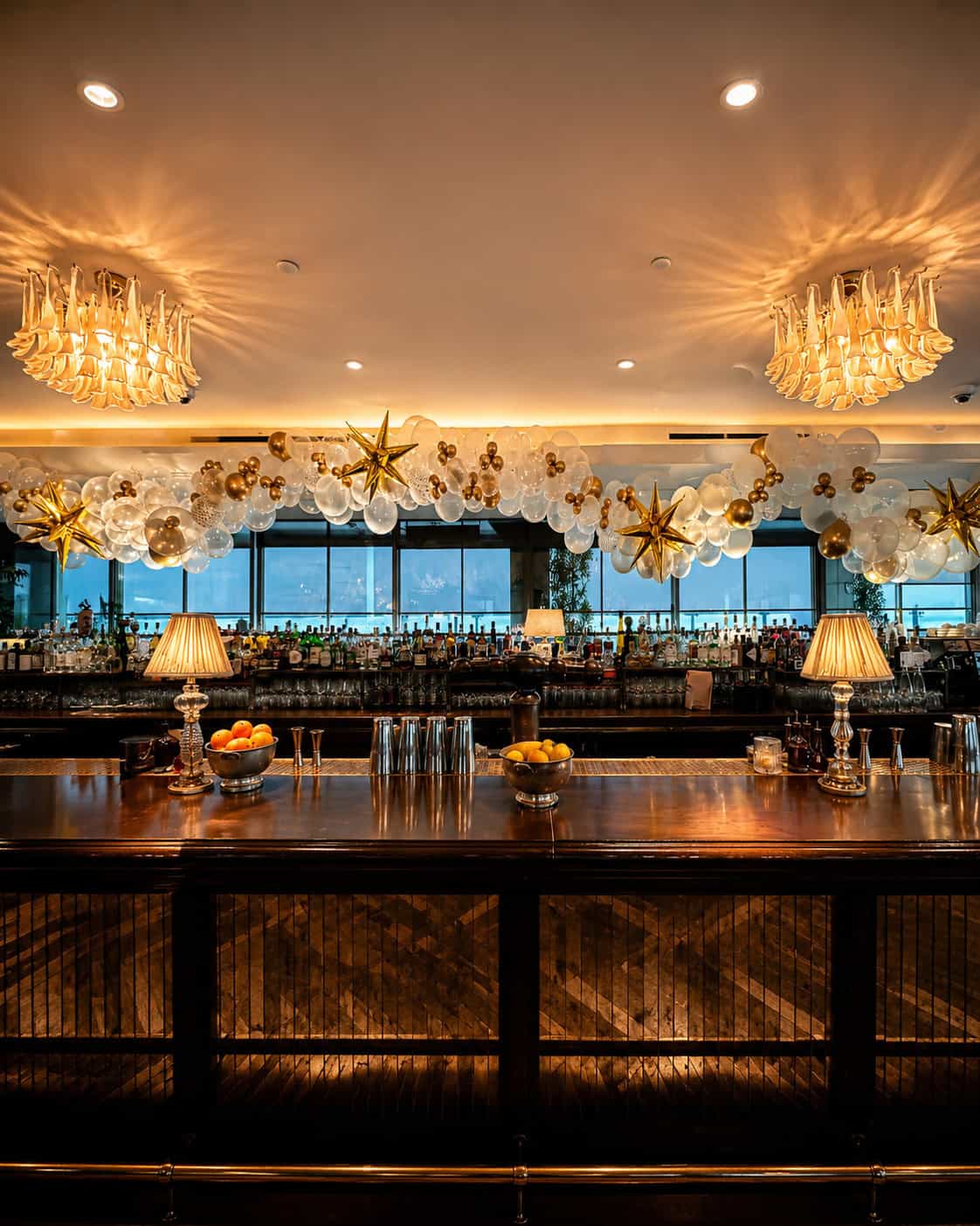 A polished bar with two lamps, bowls of citrus fruit, glassware, and festive gold and white balloon decorations creates a vibrant setting perfect for Grand Openings above shelves of bottles.