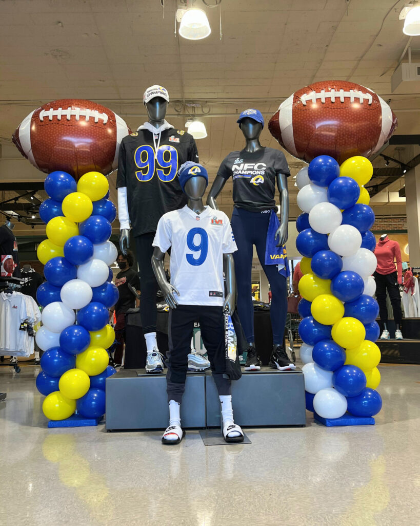 Three mannequins in Rams football apparel stand between two balloon columns with large football balloons on top, creating a lively Sports Events vibe inside the retail store.