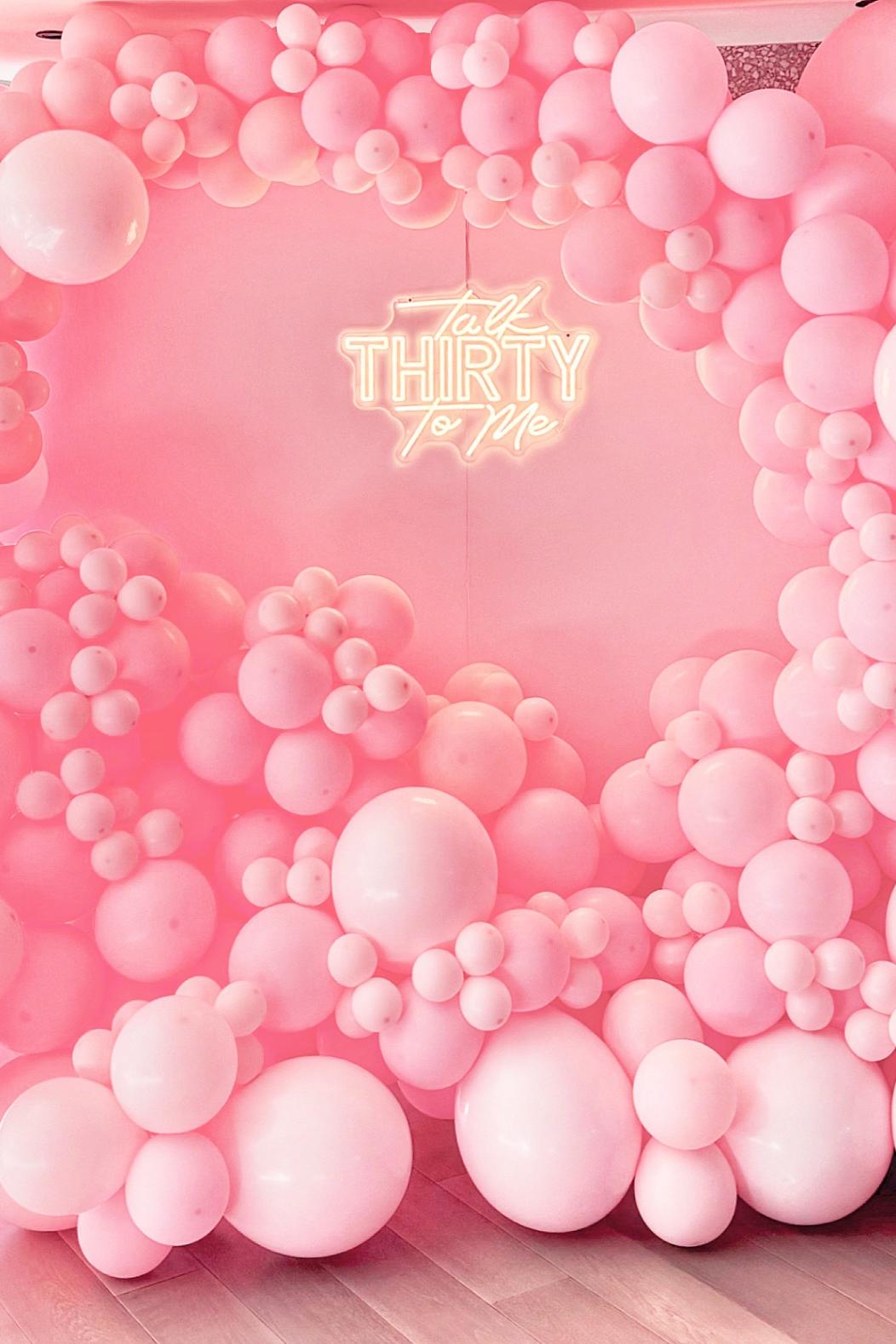 A pink balloon arch surrounds a neon sign that reads "talk THIRTY to me" against a pink wall and floor backdrop.