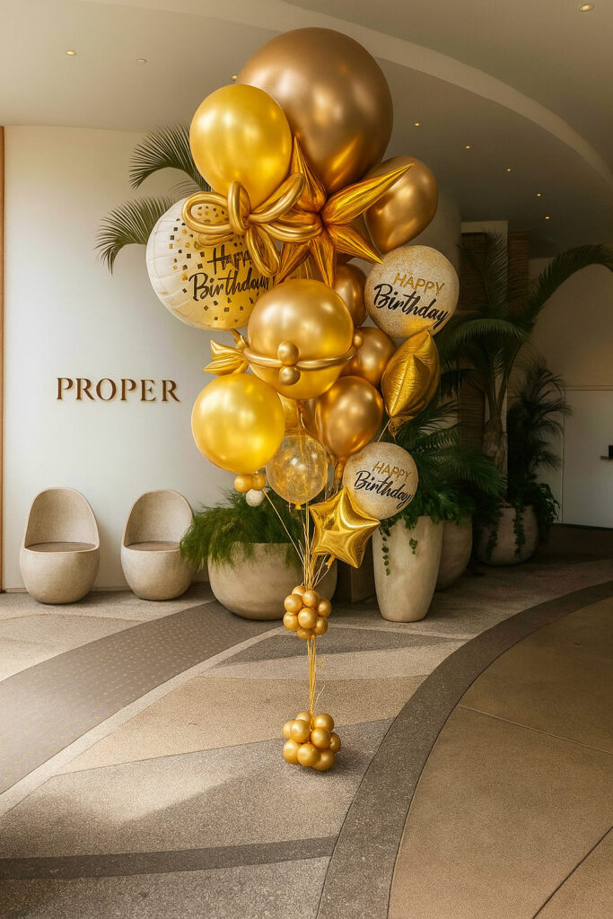A large bouquet of gold and white balloons, some with "Happy Birthday" text, arranged in a modern lobby near chairs and green plants.