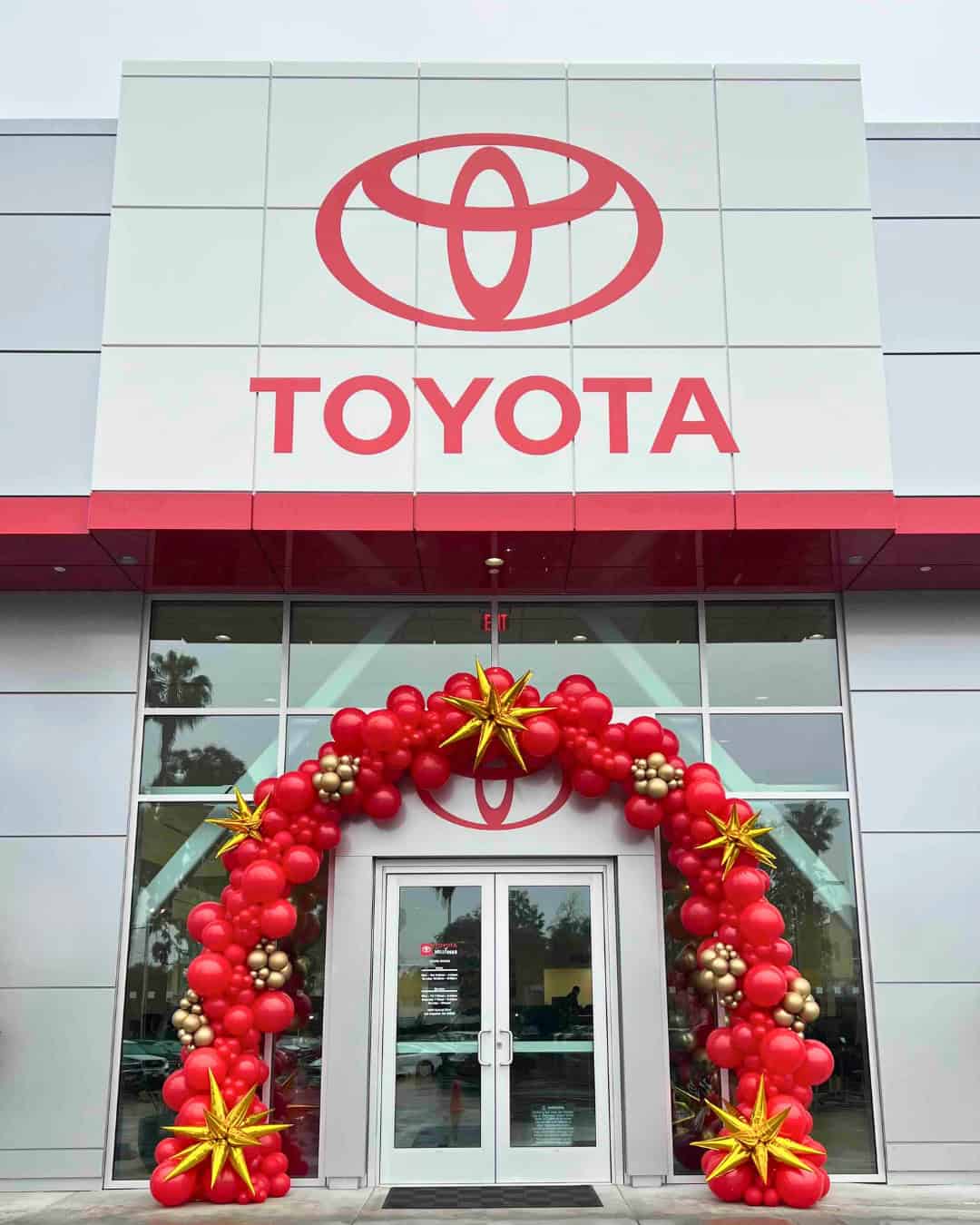 Entrance of a Toyota dealership decorated with a red and gold balloon arch, featuring large gold star-shaped balloons—perfect for Grand Openings.