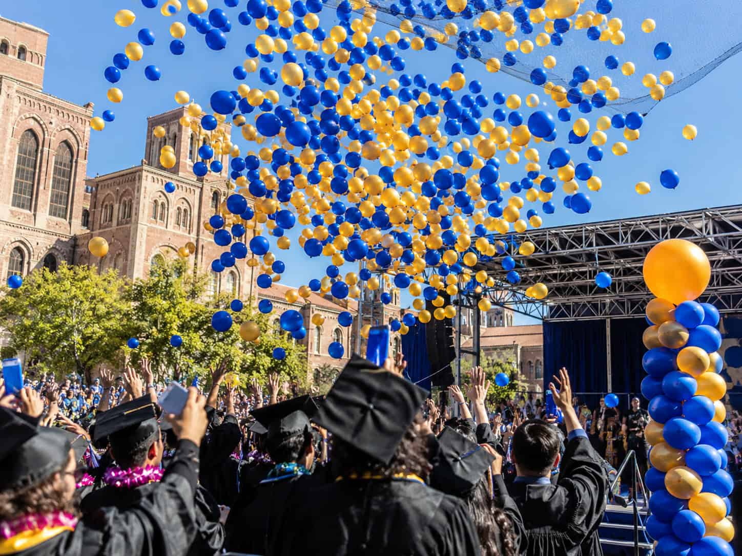 Graduates in caps and gowns celebrate outdoors as blue and yellow balloons are released into the sky during a commencement ceremony.