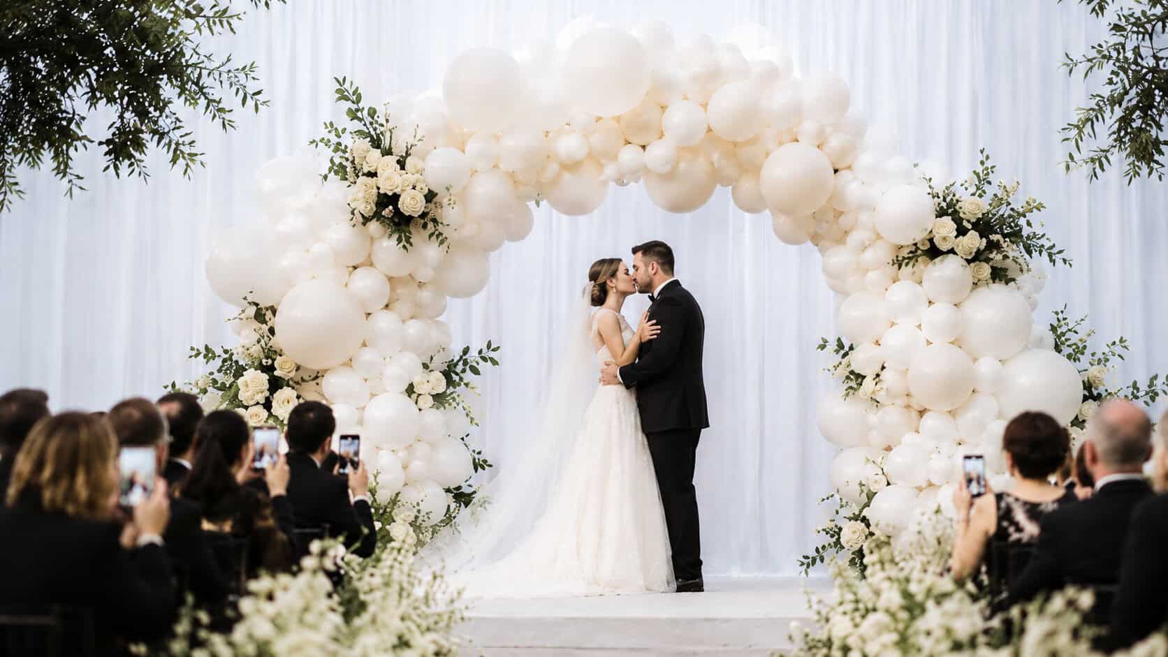 A bride and groom kiss under a large white balloon arch at their wedding ceremony, surrounded by seated guests taking photos.
