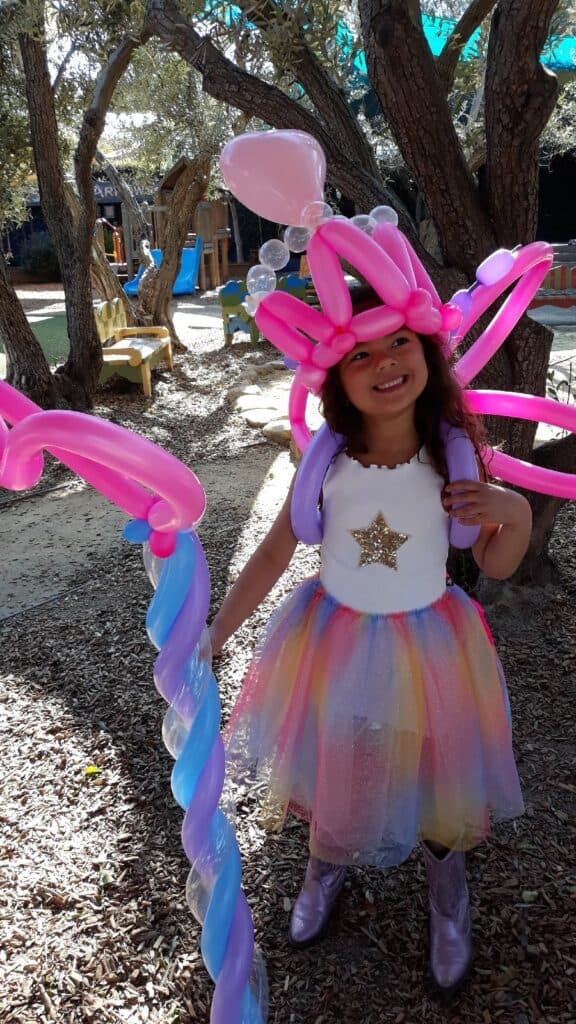 Birthday Party Tips from an LA Celebrity Party Planner 2 A young girl in a rainbow dress and boots stands outdoors, smiling and holding a large twisted balloon staff, created by a talented balloon artist, while wearing a headpiece made of pink and purple balloons.