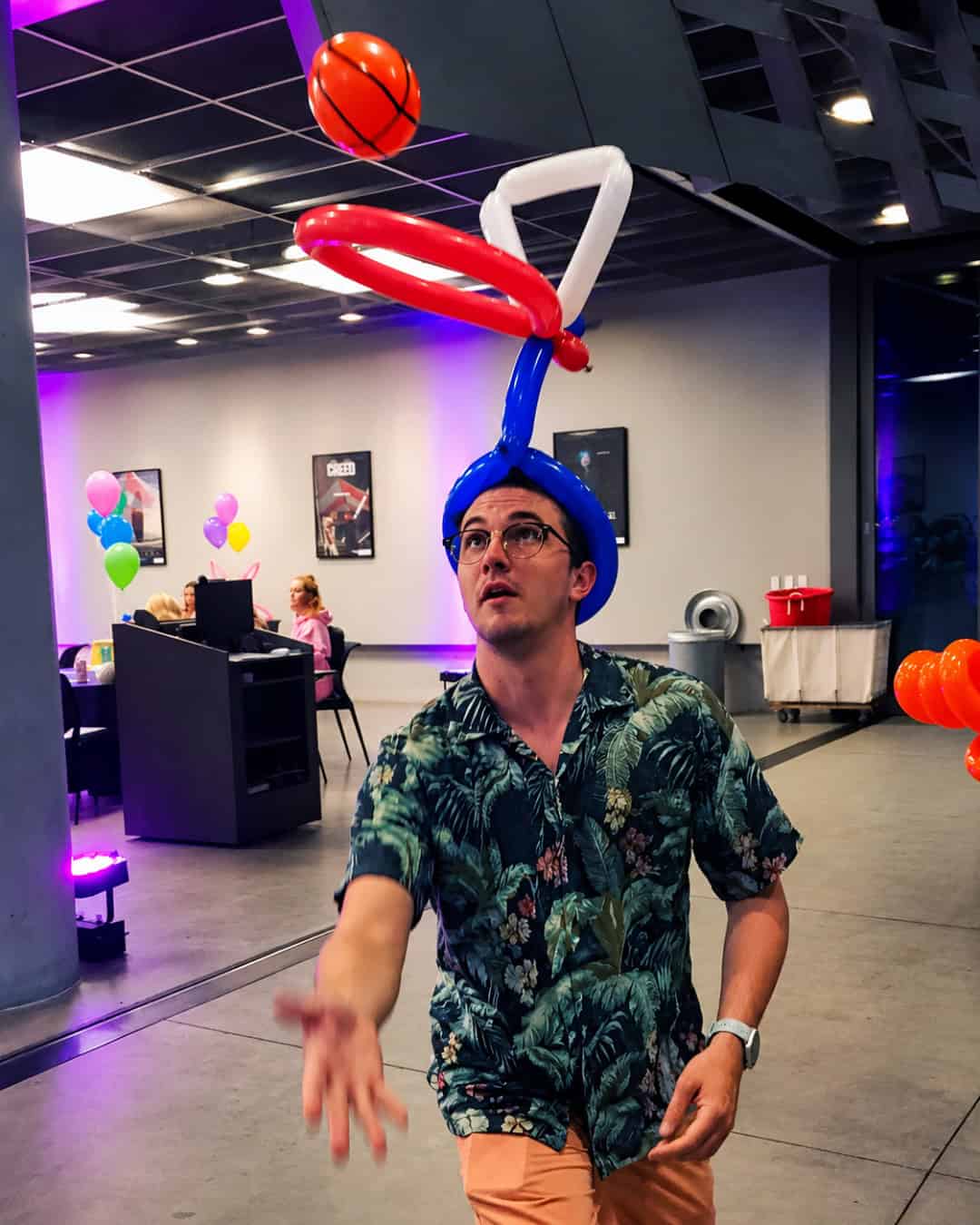 A man wearing a balloon hat shaped like a basketball hoop tosses a small basketball indoors at a decorated sports events space.