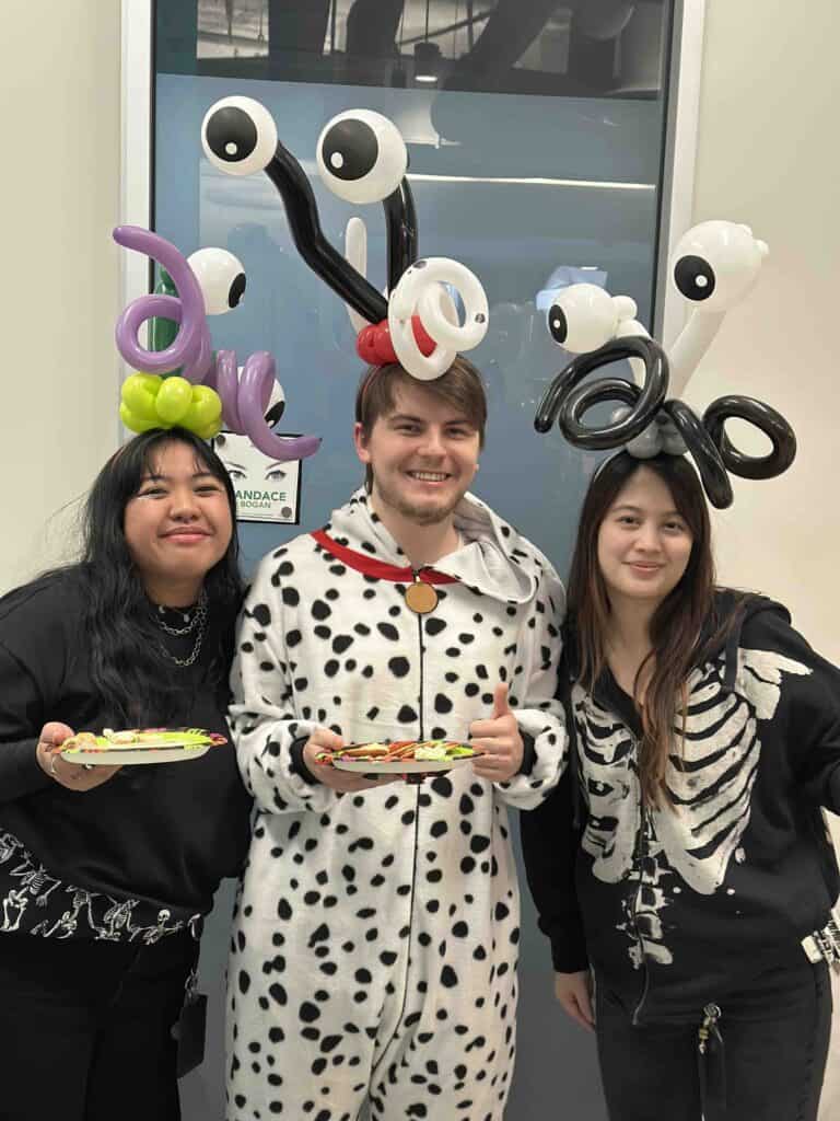 Three people pose indoors; two wear balloon twisting hats with eye designs, and one is dressed in a Dalmatian costume. All hold plates with food and smile at the camera.