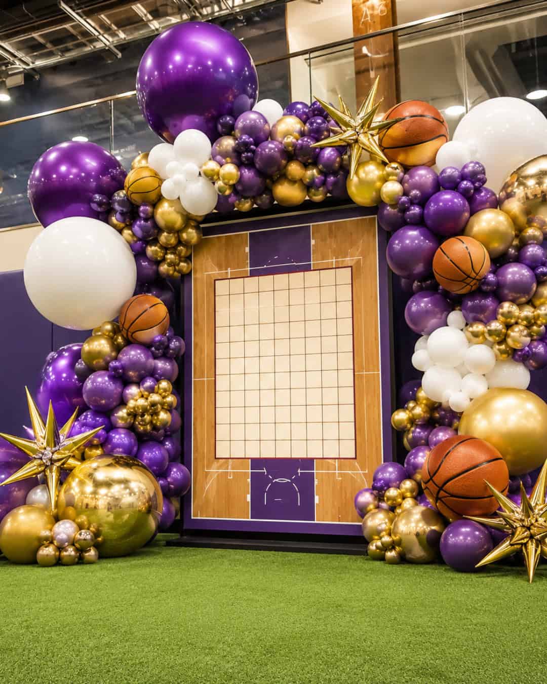 A basketball-themed photo frame decorated with purple, white, and gold balloons, gold stars, and basketballs, set on artificial grass indoors—perfect for capturing memories at sports events.