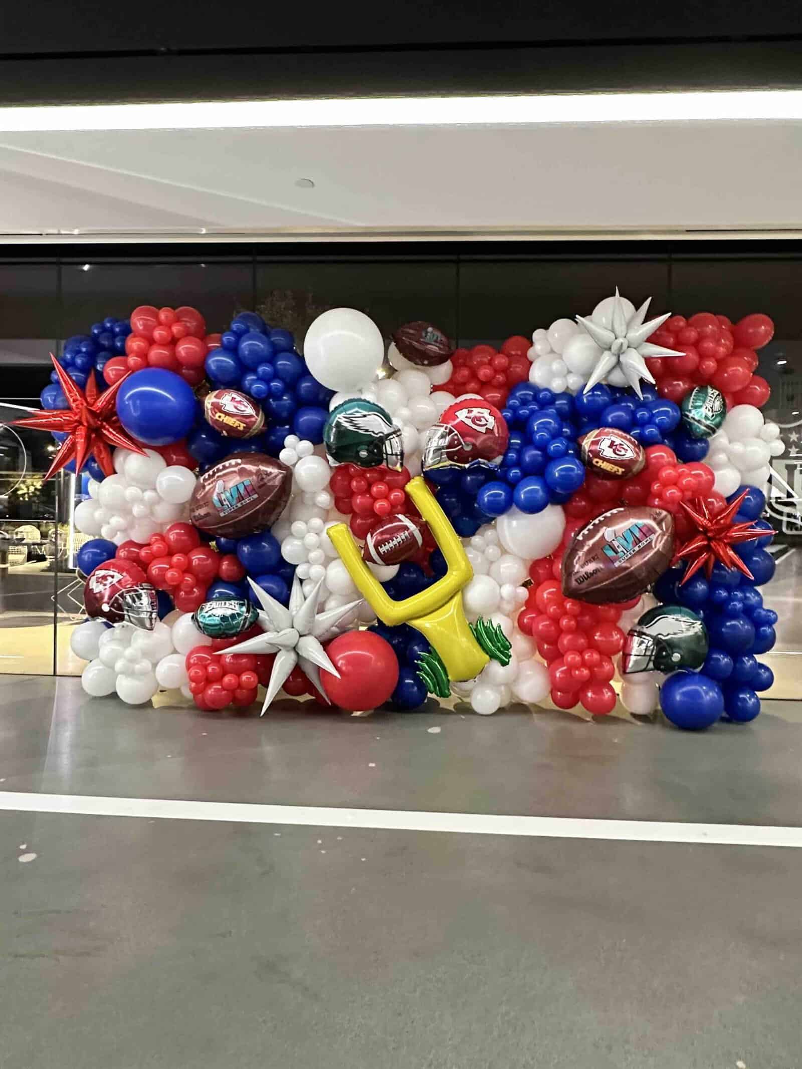 Perfect for sports events, this balloon display features red, white, and blue balloons, football balloon decorations, metallic stars, and a yellow goal post balloon set up in front of a window.