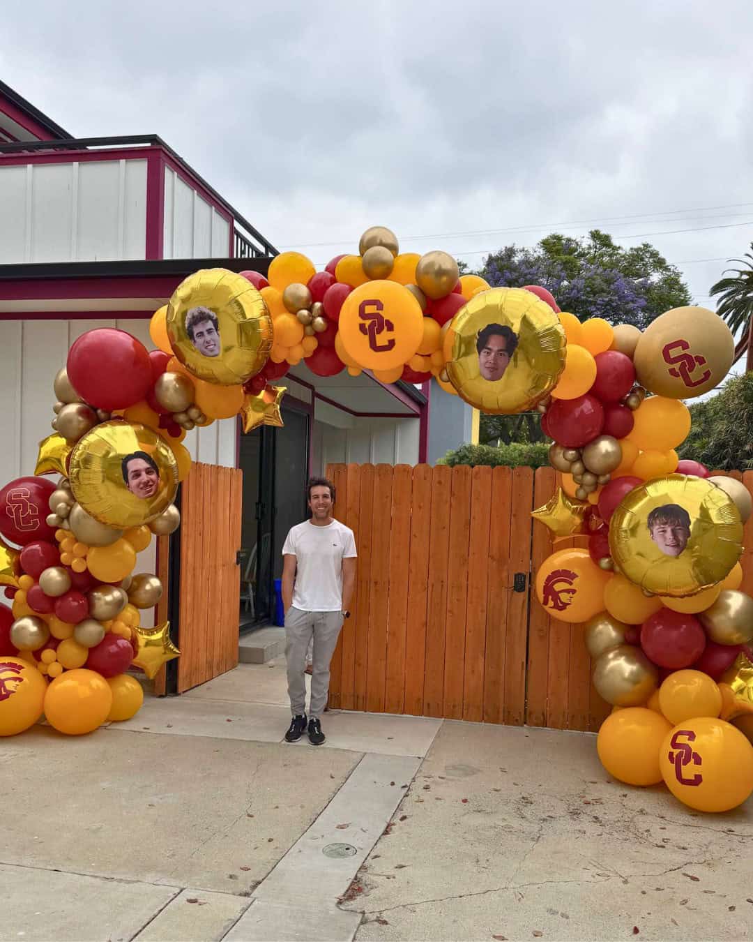 A man stands in front of a balloon arch decorated with USC logos, large balloons featuring four printed faces, and festive touches perfect for Sports Events. The setting is an outdoor area with a wooden fence and building.