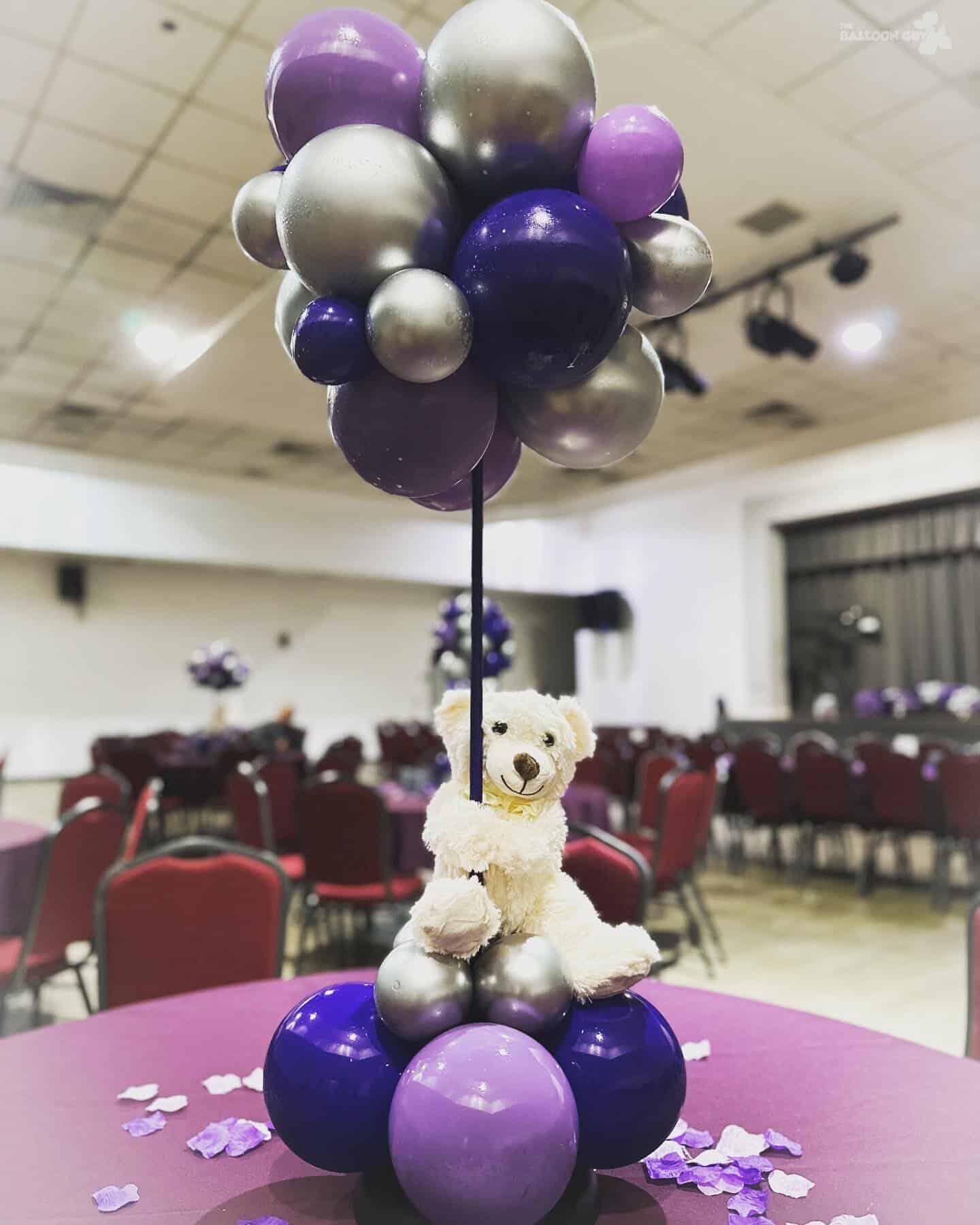 A centerpiece with purple and silver balloons, perfect for baby showers, features a teddy bear sitting at the base, set on a table in an event hall with red chairs and scattered purple petals.