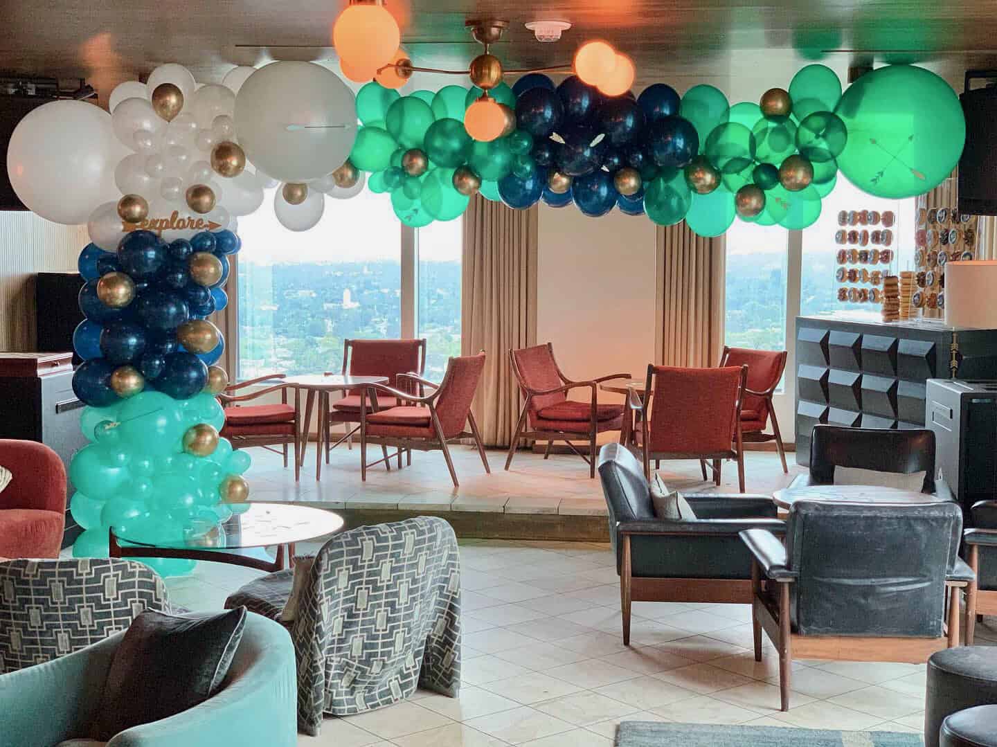 A lounge area decorated with blue, green, white, and gold balloons—perfect for baby showers—featuring chairs, tables, large windows, and a stunning city view in the background.