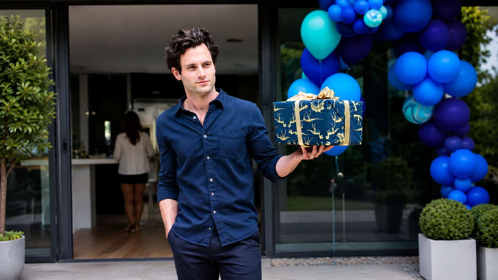 A man in a navy shirt holds a wrapped gift with a gold bow outside a building decorated with blue balloons, creating a festive scene reminiscent of TV & Film balloon decor.