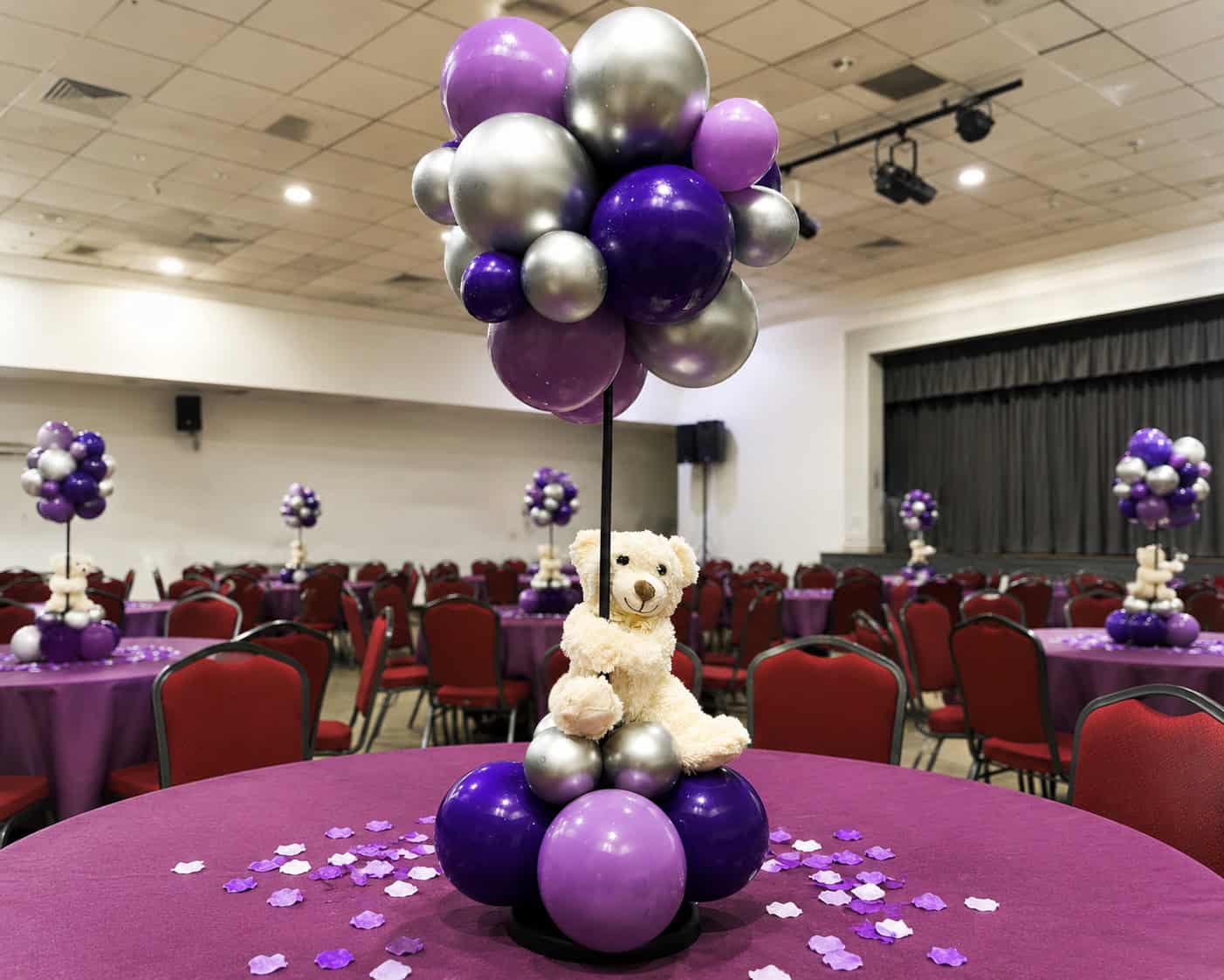 A table centerpiece perfect for baby showers features a teddy bear sitting among purple and silver balloons in a decorated event hall with matching tables and chairs.
