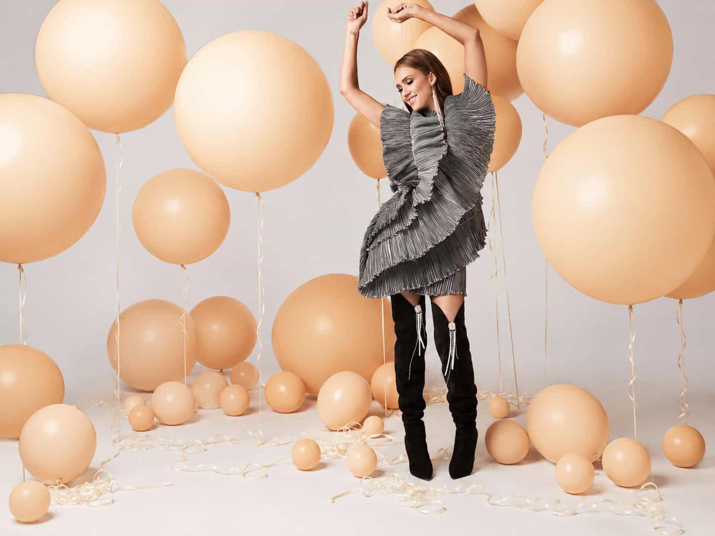 A woman in a ruffled grey dress and black boots poses with arms raised among large beige balloons on a studio set, creating an atmosphere reminiscent of TV & Film balloon decor.