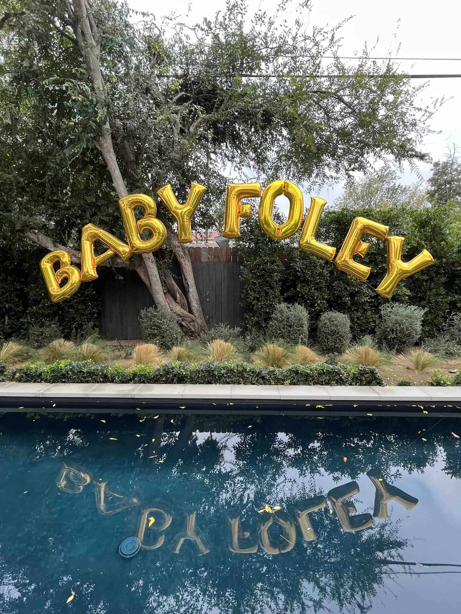 Gold balloons spelling "BABY FOLEY" float above a backyard pool, their shimmering letters and surrounding trees beautifully reflected in the water—perfect decor for unforgettable baby showers.