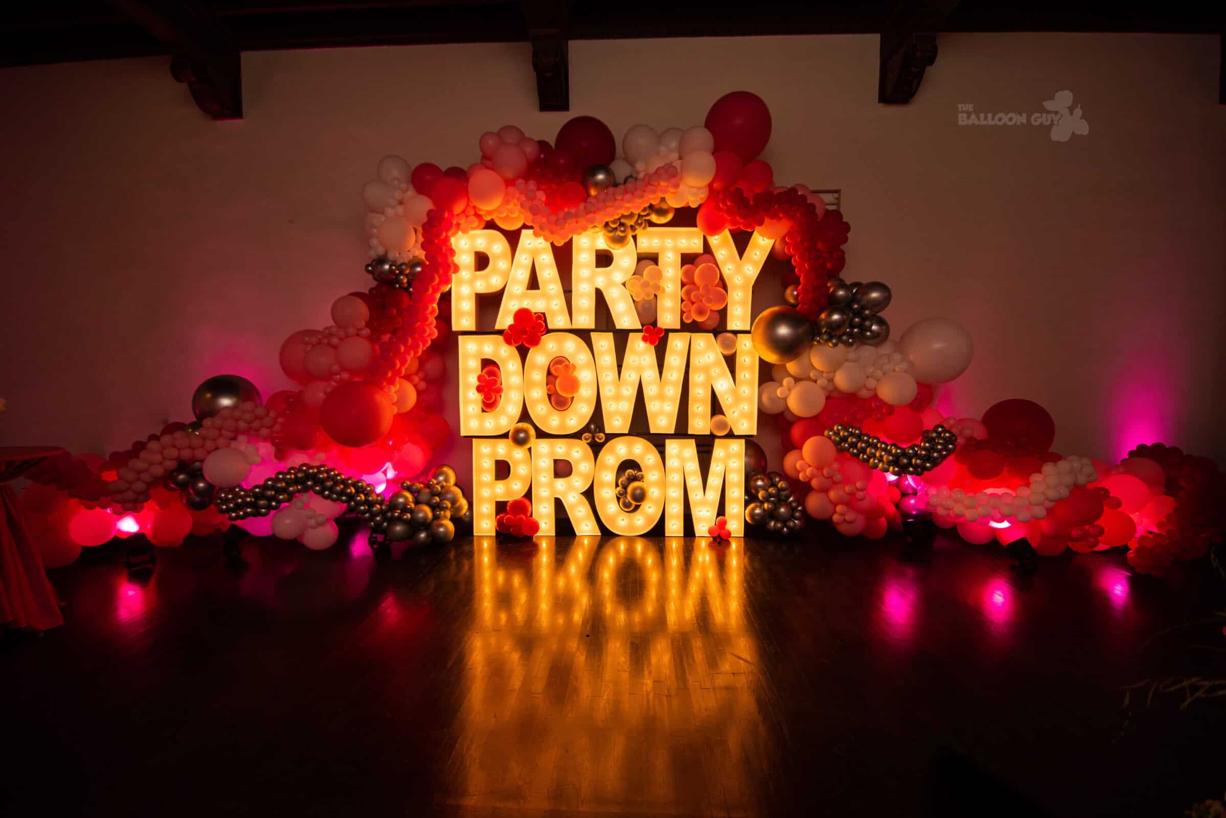 Large illuminated letters spelling "PARTY DOWN PROM" are surrounded by red, white, pink, and silver balloons against a dimly lit wall, creating red carpet event balloon decor vibes.