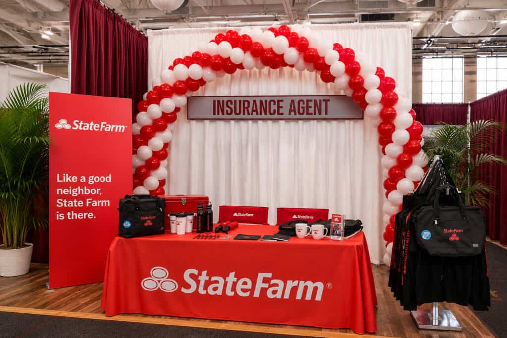 A State Farm insurance agent booth with TV & Film balloon decor featuring a red and white balloon arch, branded table, promotional items, and banners displaying the State Farm logo and slogan.