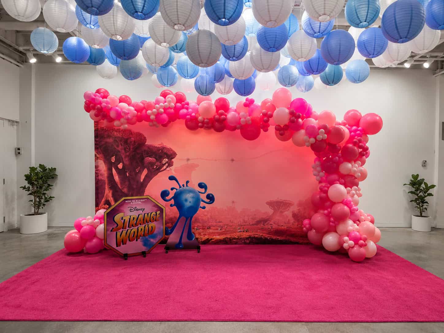 Photo of a Disney Strange World backdrop with pink balloons, a blue character cutout, and a pink carpet. Blue and white paper lanterns hang from the ceiling, creating stylish red carpet event balloon decor.