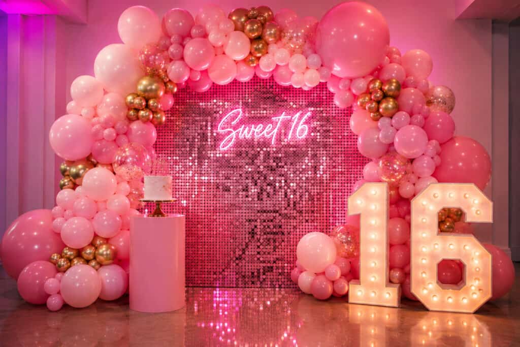 A pink and gold sweet 16 balloon decor arch frames a sequin backdrop with a neon "Sweet 16" sign, a cake on a pink stand, and large illuminated numbers "16".