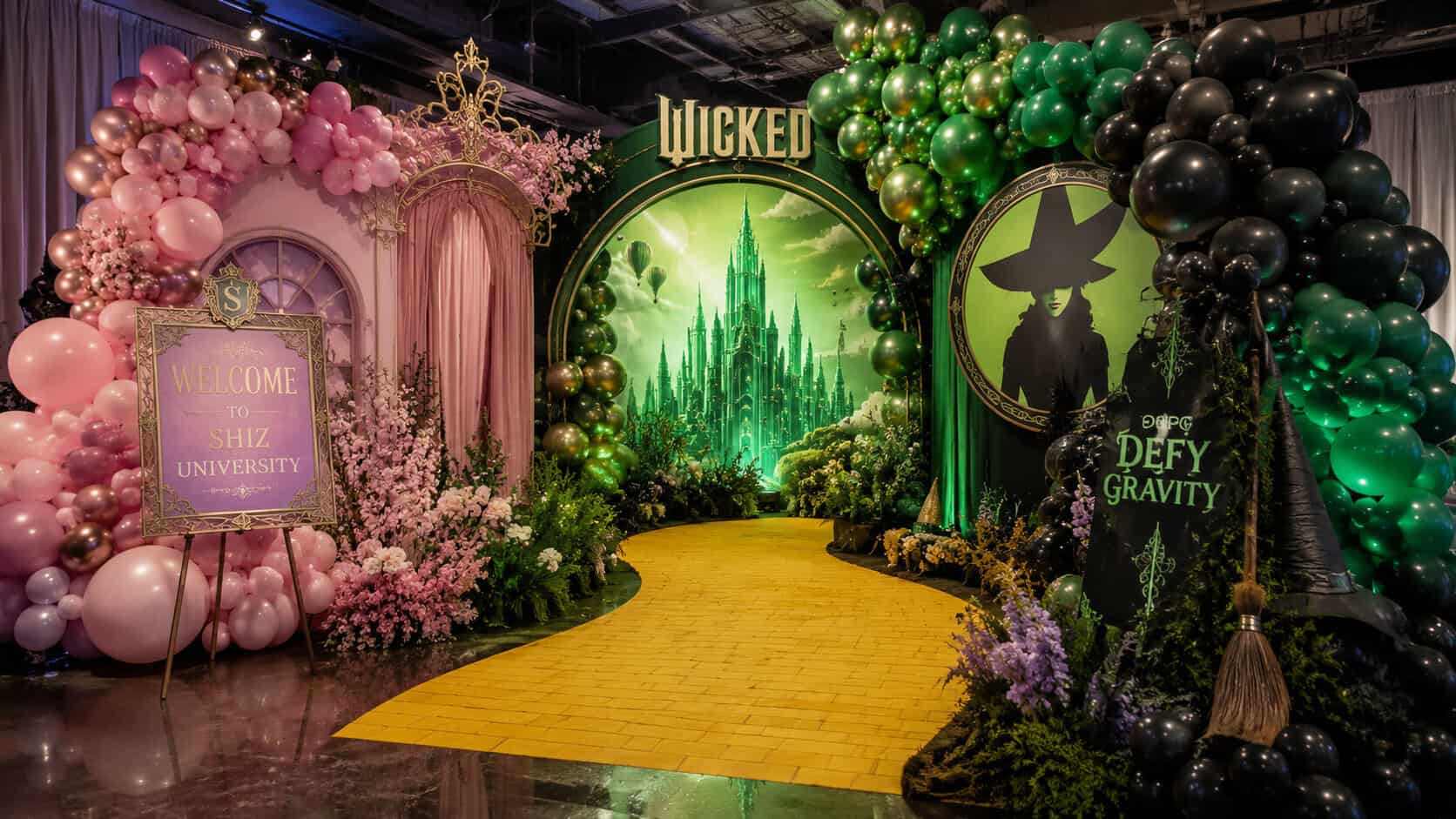 A "Wicked" themed display featuring a yellow brick road, green and pink TV & Film balloon decor, and signs reading "Welcome to Shiz University" and "Defy Gravity," with an Oz backdrop.