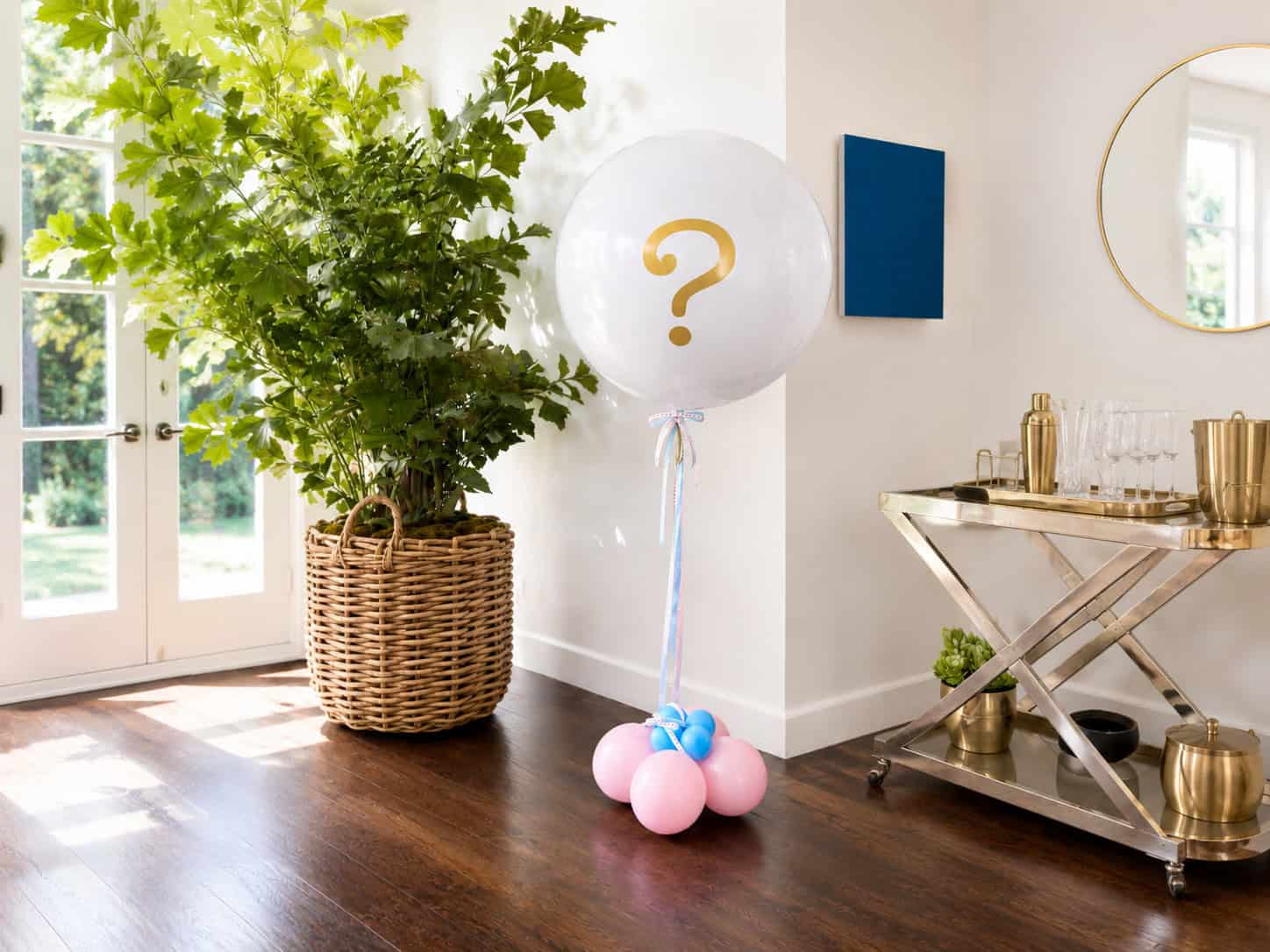 A large white balloon with a gold question mark, perfect for a Gender Reveal, stands on pink and blue smaller balloons in a bright room with a plant and a bar cart.