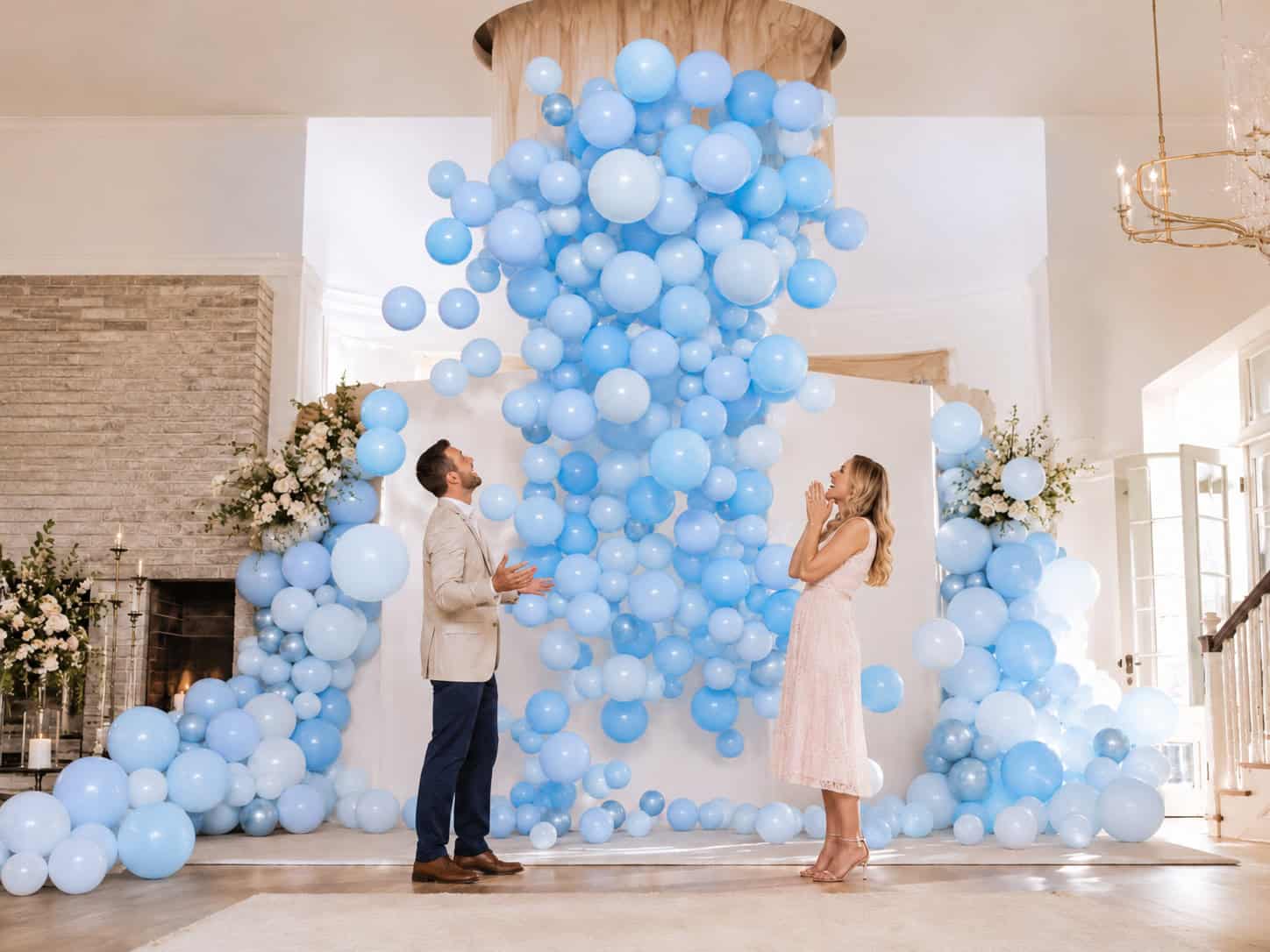 A man and woman stand facing each other indoors at a gender reveal, surrounded by numerous light blue balloons cascading from the ceiling in front of a white backdrop.