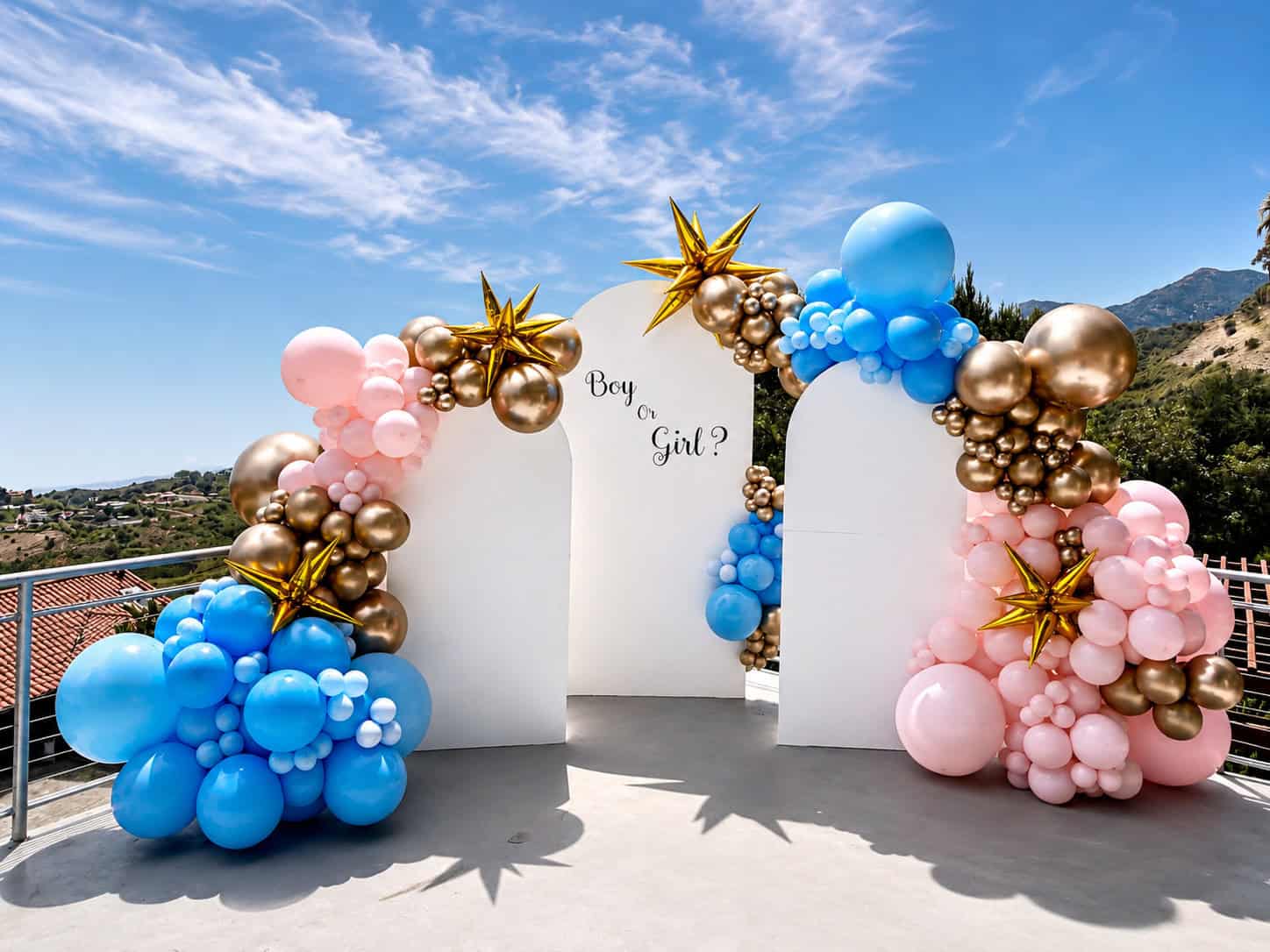 Balloon arch with blue, pink, gold, and star-shaped balloons surrounds a white panel reading "Boy or Girl?"—perfect for a Gender Reveal on an outdoor terrace under a blue sky.