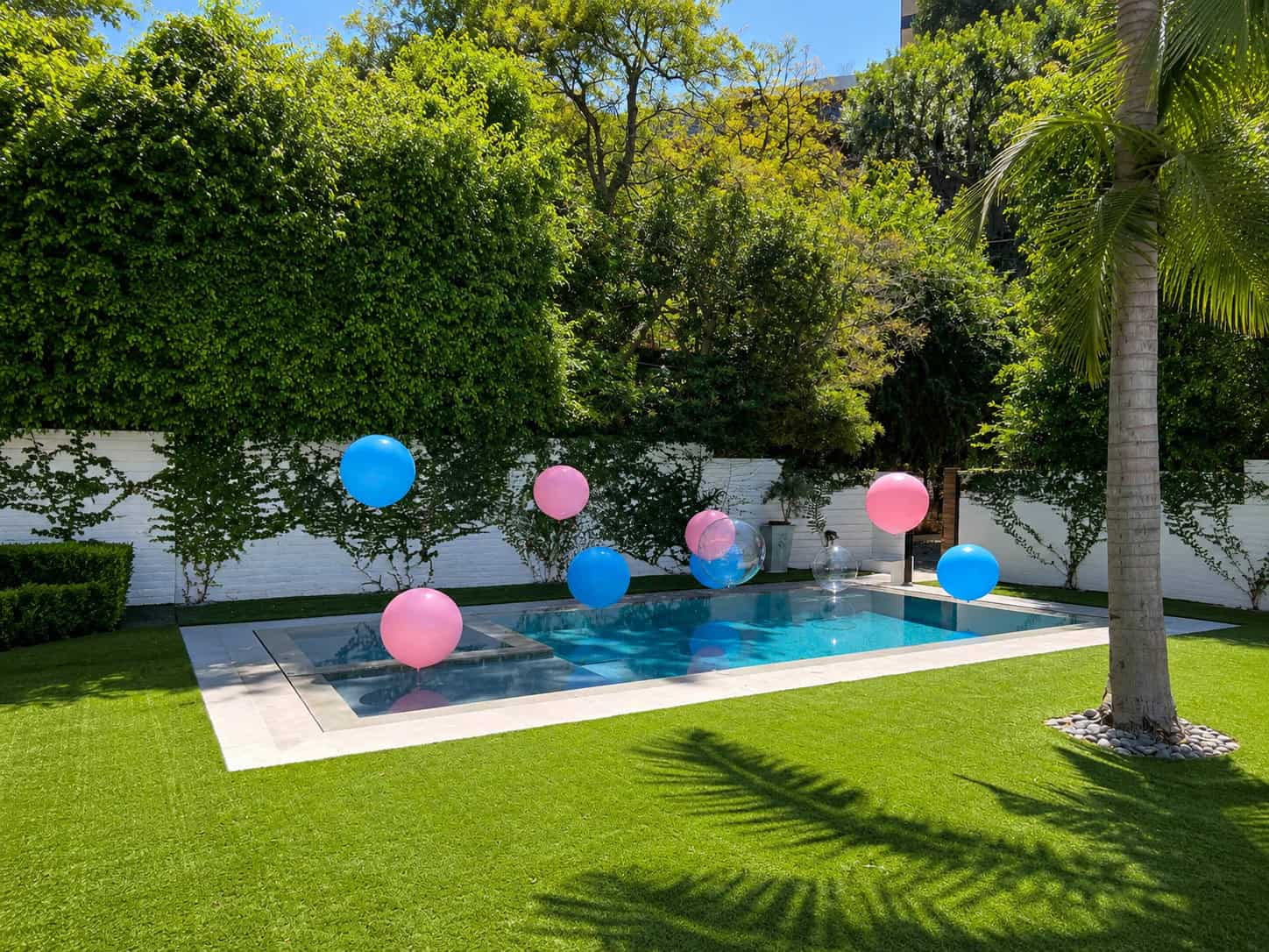 A small backyard swimming pool is surrounded by artificial grass and greenery, with pink and blue balloons floating above the water for a festive gender reveal.