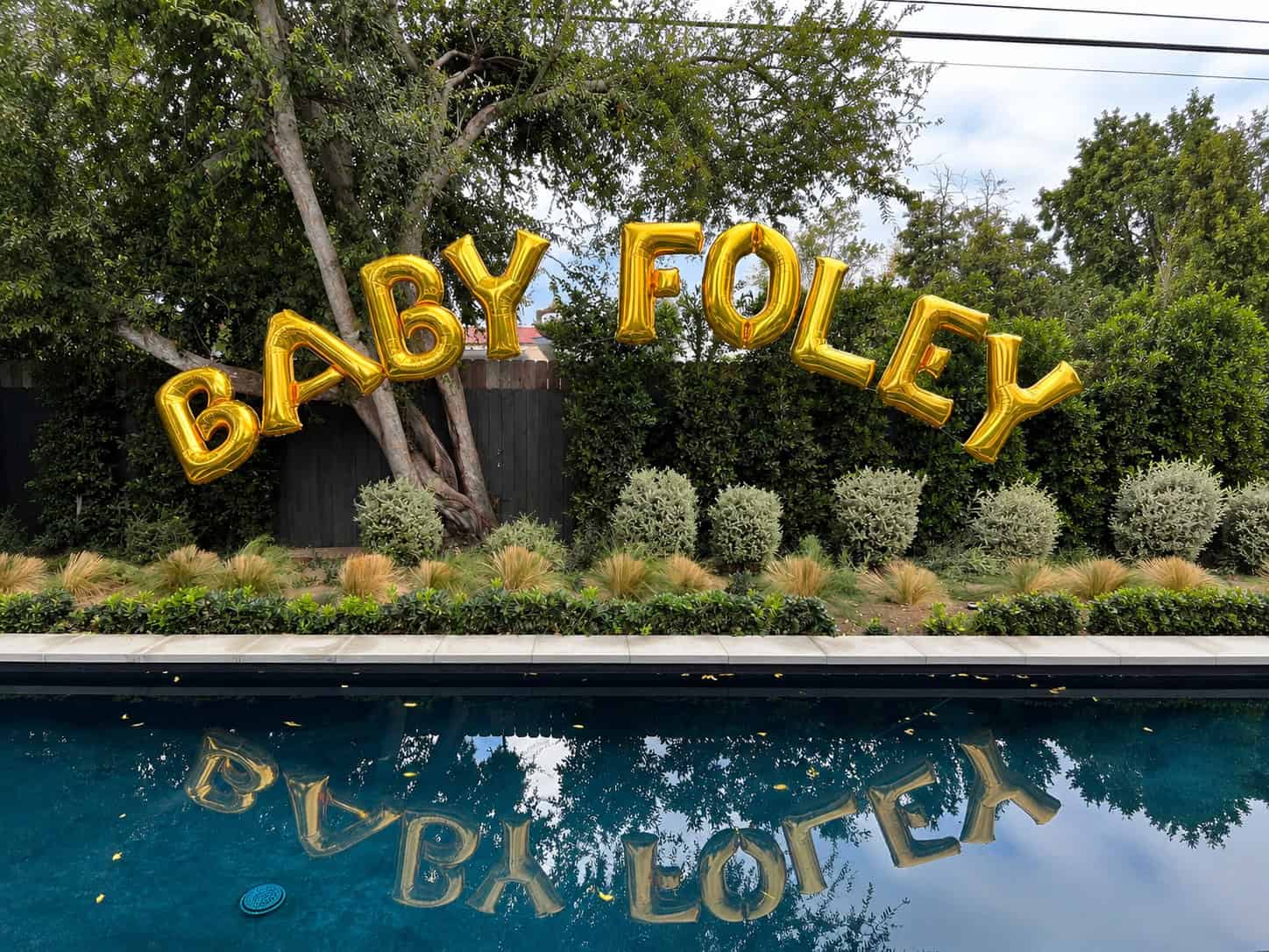 Gold letter balloons spelling "BABY FOLEY" are displayed by a backyard pool, their reflection shimmering in the water—perfect décor for stylish baby showers.