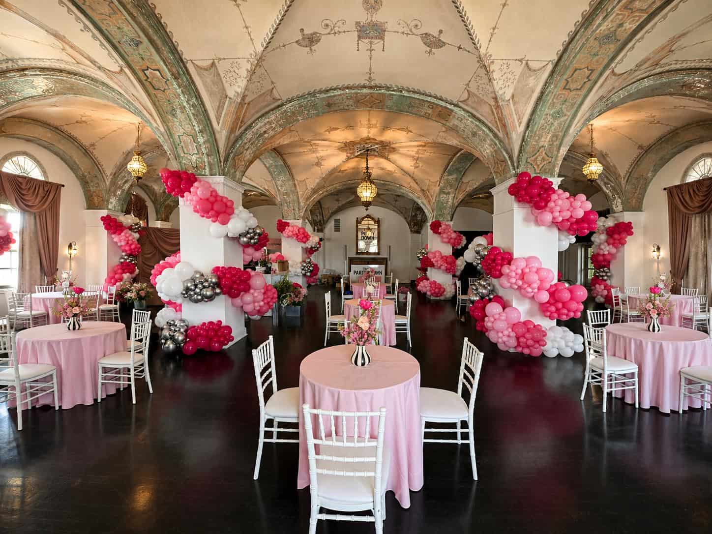 Elegant banquet hall decorated with clusters of pink, white, and silver balloons, perfect for baby showers. Round tables with pink tablecloths, white chairs, and ornate vaulted ceilings complete the charming setting.