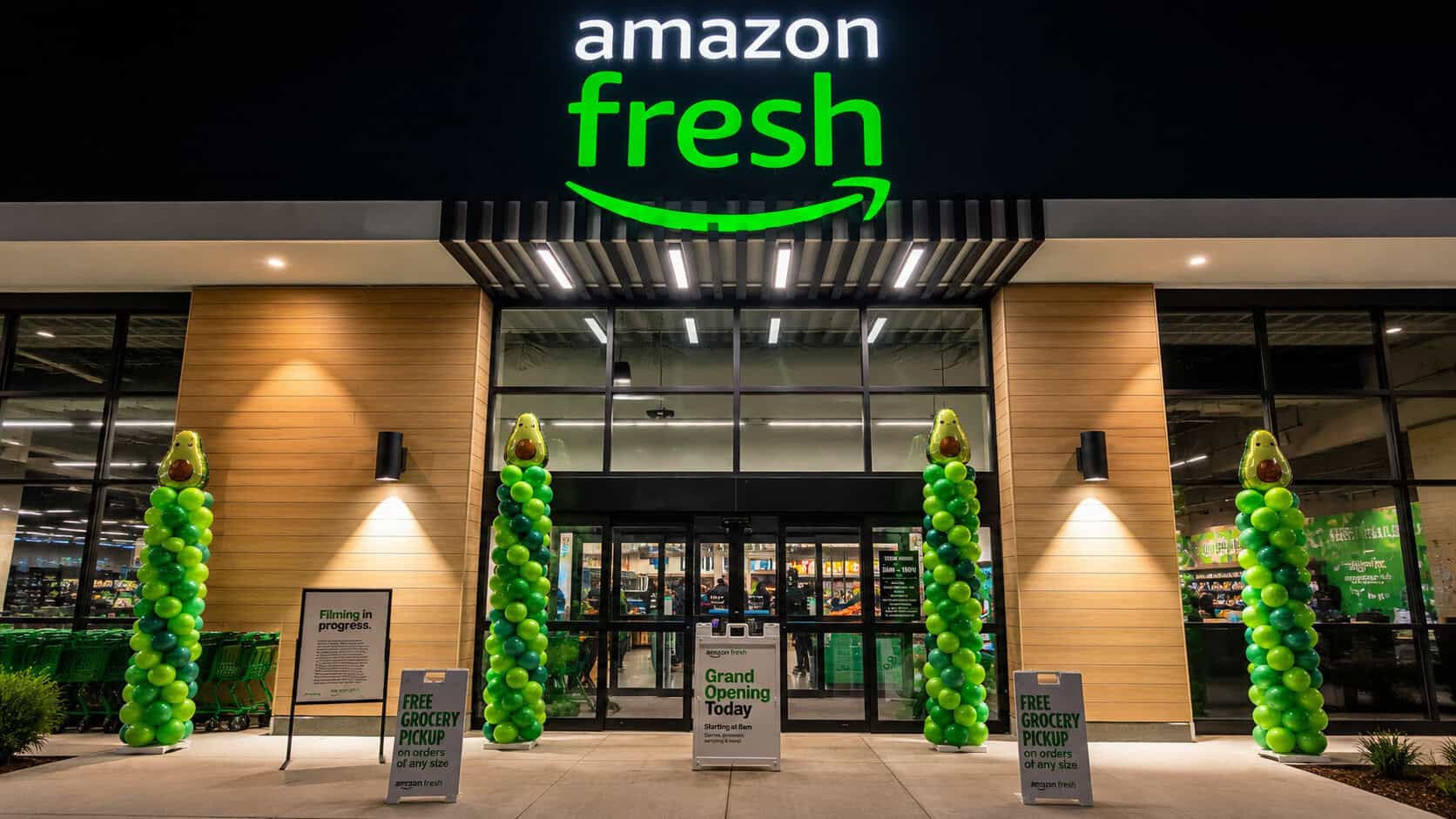 Amazon Fresh grocery store exterior at night with custom balloon columns in vibrant green and grand opening signs welcoming customers at the entrance.