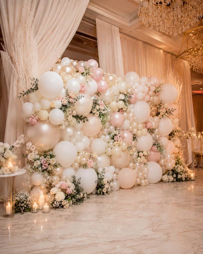 Balloon and floral backdrop with a stunning balloon wall featuring white, cream, and blush balloons, fresh flowers, candles, and draped curtains in an elegant, chandelier-lit setting.