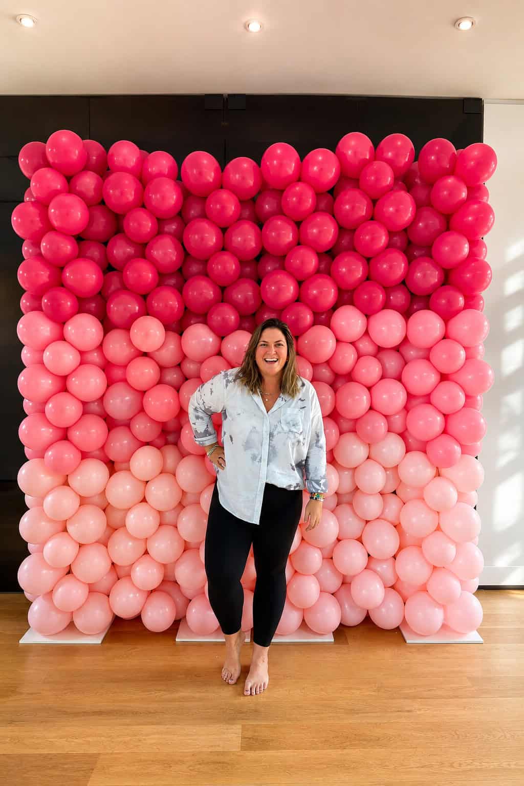 A woman stands barefoot in front of a vibrant pink ombre balloon wall, smiling with one hand on her hip.