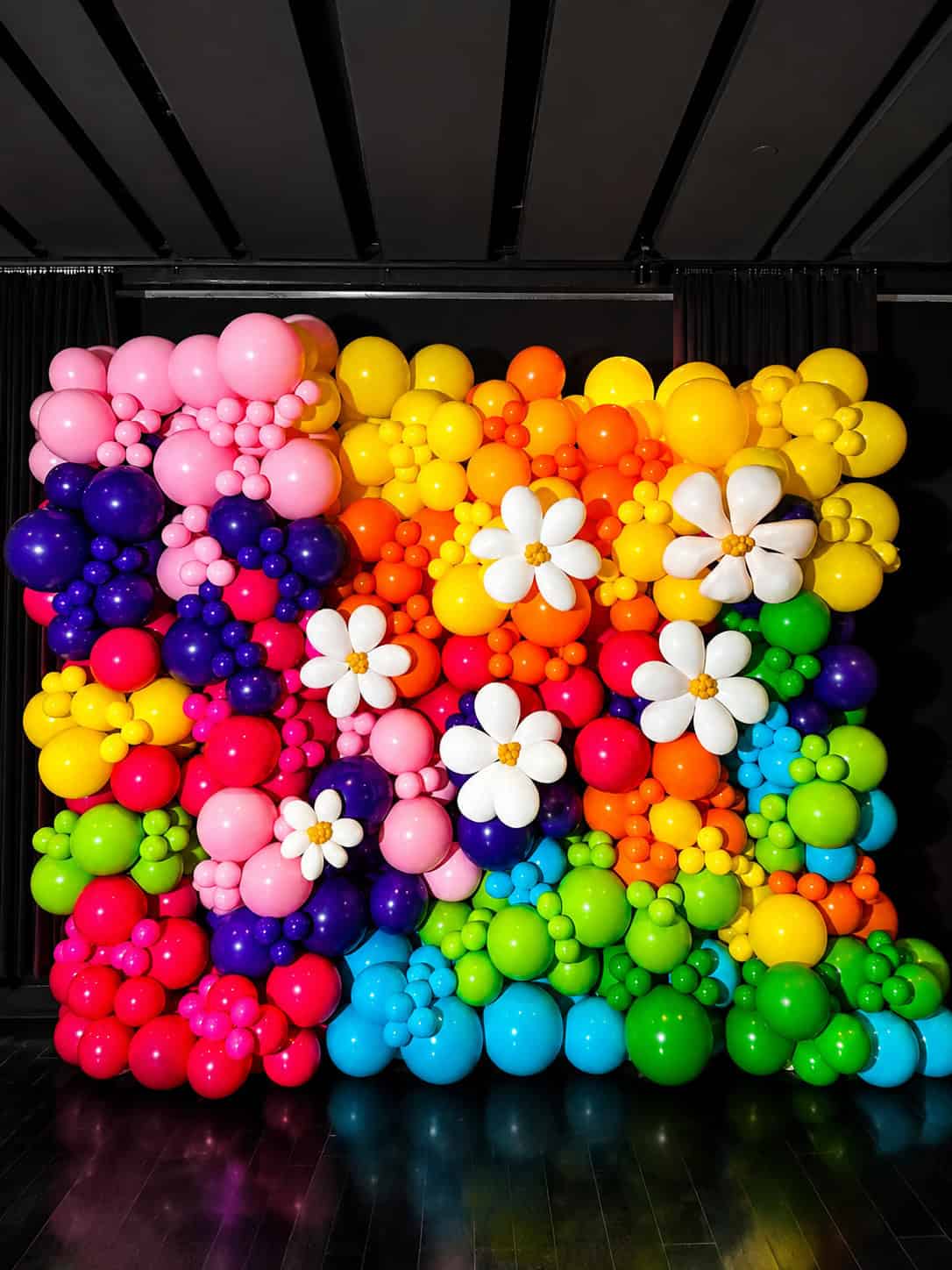 A vibrant balloon wall arranged in a grid pattern showcases clusters of balloons in pink, purple, blue, green, yellow, orange, and red, beautifully decorated with large white balloon flowers.