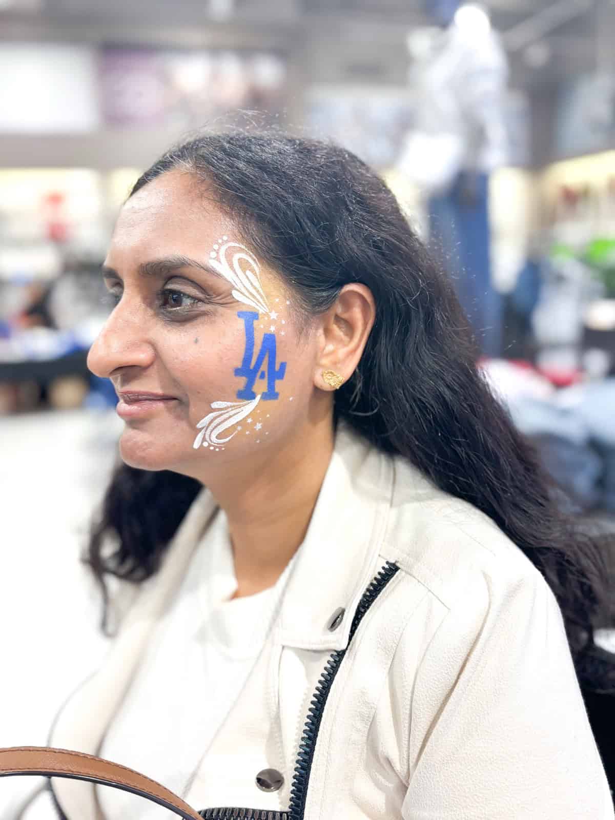 Woman with long dark hair and a white jacket has an LA Dodgers logo and white decorative face painting on her left cheek; background shows a blurred indoor setting.