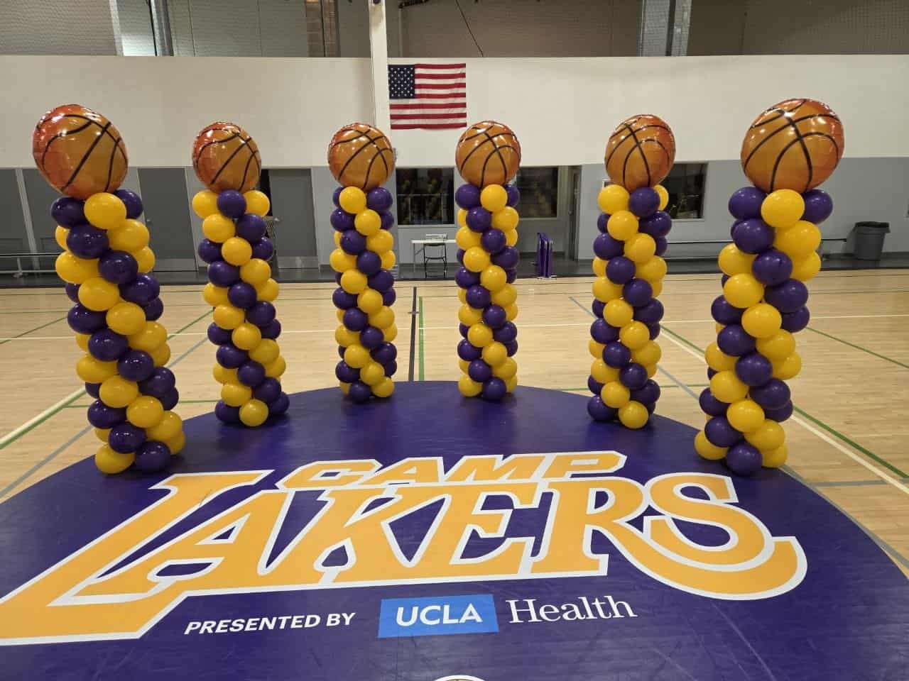 Six custom balloon columns with basketball toppers stand on a gym floor featuring a large “Camp Lakers” logo, with an American flag in the background.