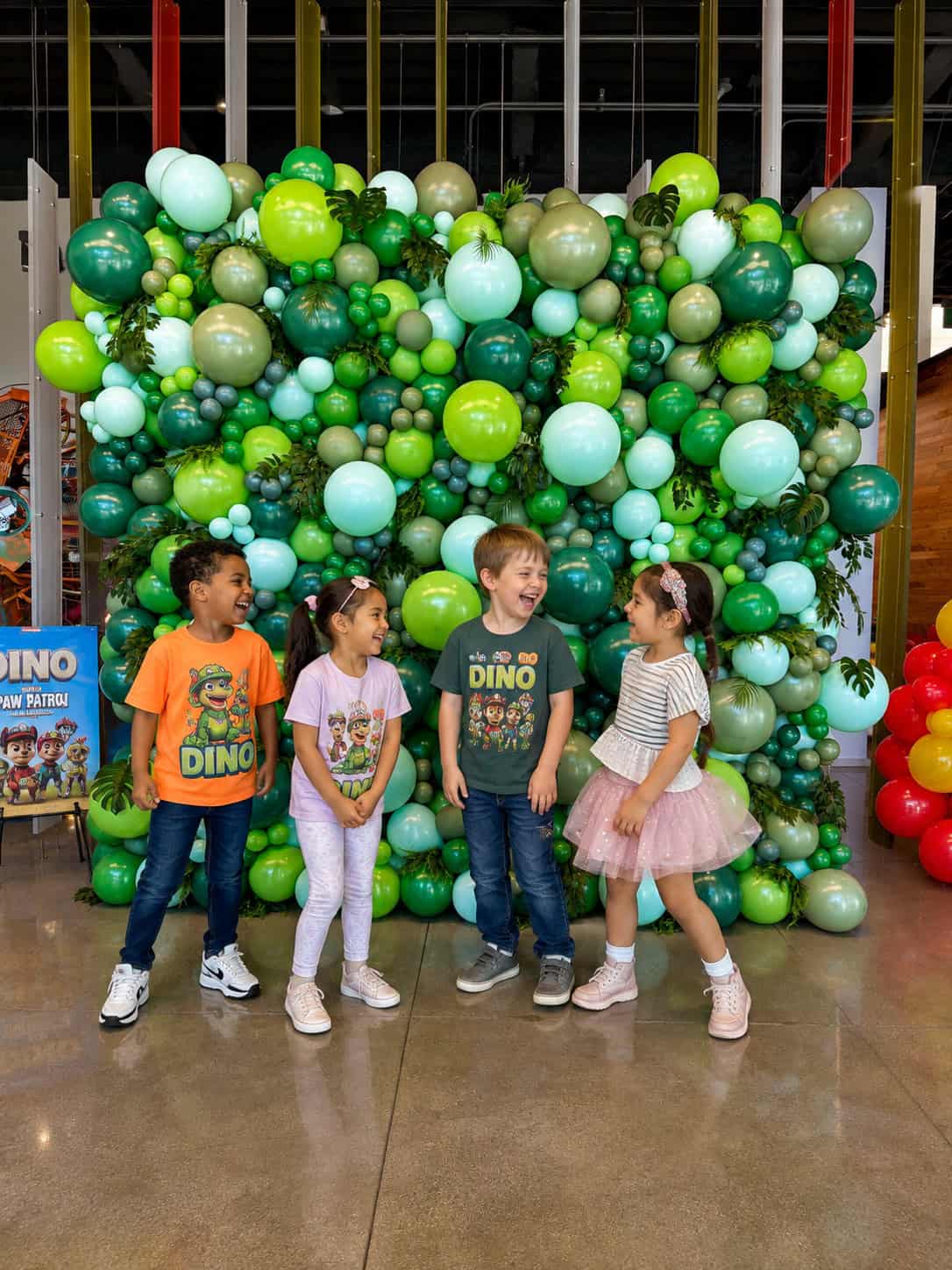 Four children stand smiling in front of a vibrant balloon wall decorated with green and white balloons, wearing colorful casual clothes and fun dinosaur-themed shirts.