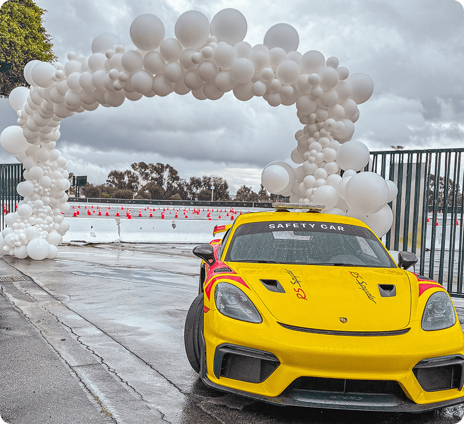A yellow safety car is parked near a wet track, beside a balloon arch made of white balloons. Orange cones and a cloudy sky are visible in the background.